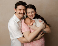 New parents embracing their sleeping newborn baby wrapped in a soft white swaddle during an intimate family newborn photography session by Kaitlyn Dawn Photography.