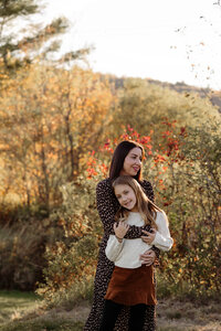A mom stands beside her daughter while the daughter gazes into the camera in a Maine home. Fine art lifestyle family photography by Damini Photography capturing genuine moments.