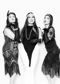 Vintage‑inspired fine art portrait from Luxe Portraiture LV featuring three women in glamorous 1920s flapper attire with sequined dresses, pearl necklaces, decorative headpieces, and confident poses, captured in black‑and‑white luxury portrait photography.