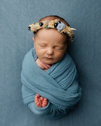 Sleeping newborn baby wrapped in a soft blue swaddle and wearing a floral headband during a studio newborn photography session with newborn photographer Kaitlyn Dawn Photography.