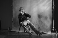 Black and white studio portrait of a man seated on a stool, backdrop and lighting stands visible in frame.