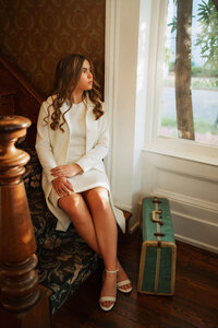 Senior portrait at the Malaga Inn in Mobile, Alabama— inside the inn, sitting on the steps next to her green suitcase looking out the window.