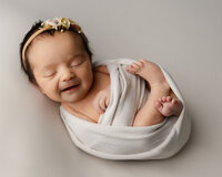 Smiling newborn baby girl wrapped in a soft white swaddle and wearing a floral headband during a studio newborn photography session in Yucaipa by Kaitlyn Dawn Photography.