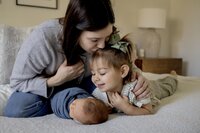A mom kisses her daughter's head as they both look at the new baby brother.