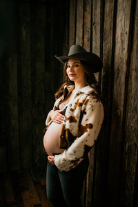 Pregnant woman in a cow-print jacket and cowboy hat, posing confidently against a rustic wooden wall during a western-style maternity session near Oconomowoc, Wisconsin.