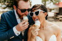 Bride and groom in sunglasses share ice cream cones during playful wedding day celebration.