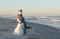 Jacksonville, FL wedding photographer capturing In St. Augustine, newlyweds share a seaside kiss as the groom playfully lifts his back foot off the sand and the bride holds her bouquet high in celebration.