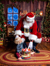 Young child gazes up at Santa while playing with a glowing carousel during a Santa's Magical Christmas photography session in Jacksonville, Florida with Nature's Child Photography, surrounded by vintage Christmas toys, twinkling holiday lights, and cozy North Pole magic.