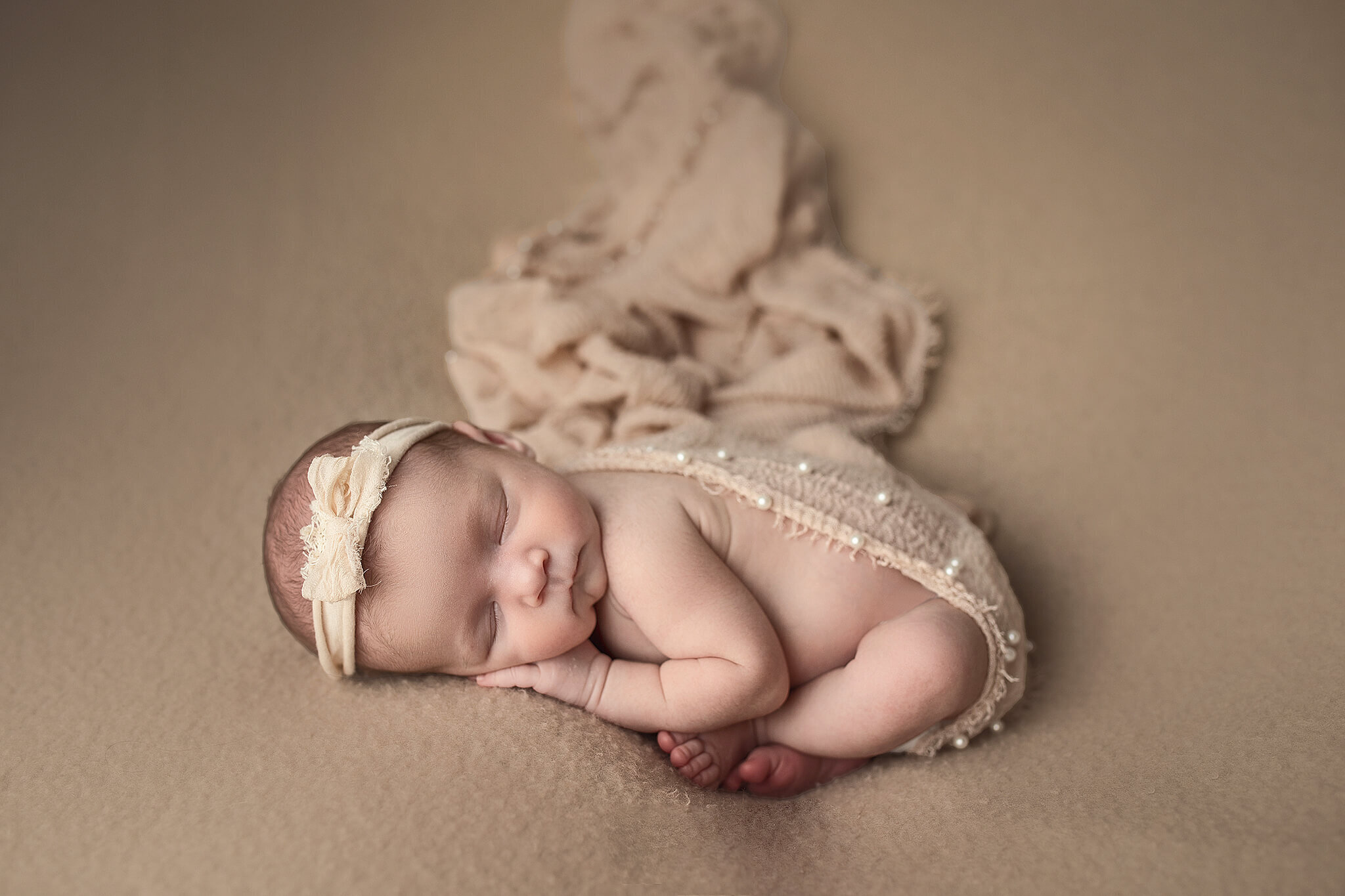 Newborn baby girl in tan wrap pose in the womb pose with tan bow on her head