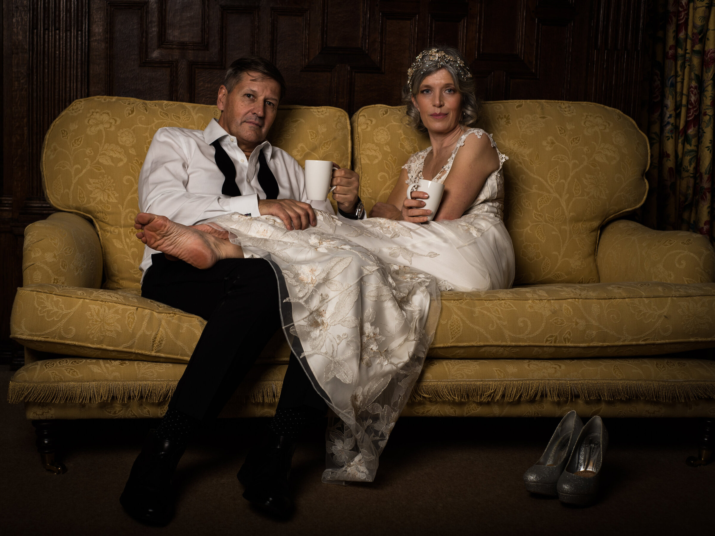 An elegantly attired couple in their 50’s share a moment of relaxed intimacy on a vintage yellow couch, sipping cups of tea and celebrating their private elopement in a setting that whispers of classic charm and quiet revelry