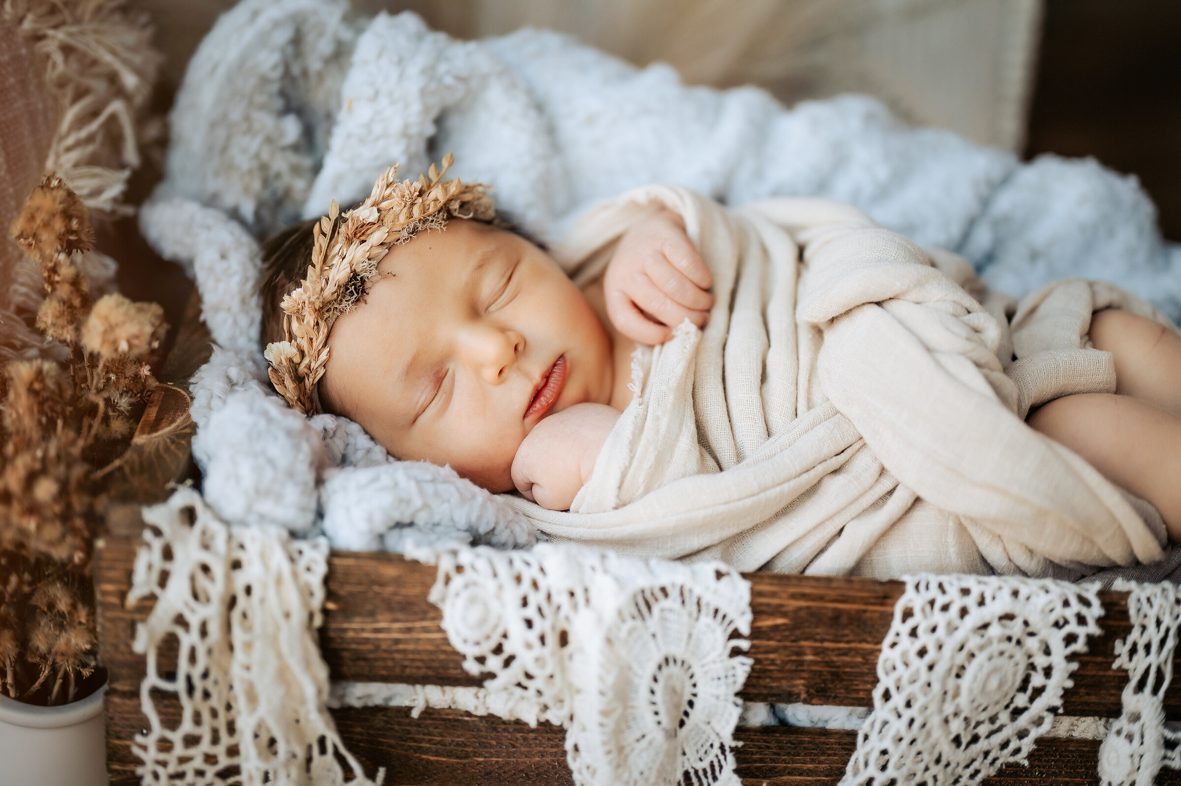 Baby lays in milk crate swaddled in linen with lace hanging over the edge of the basket