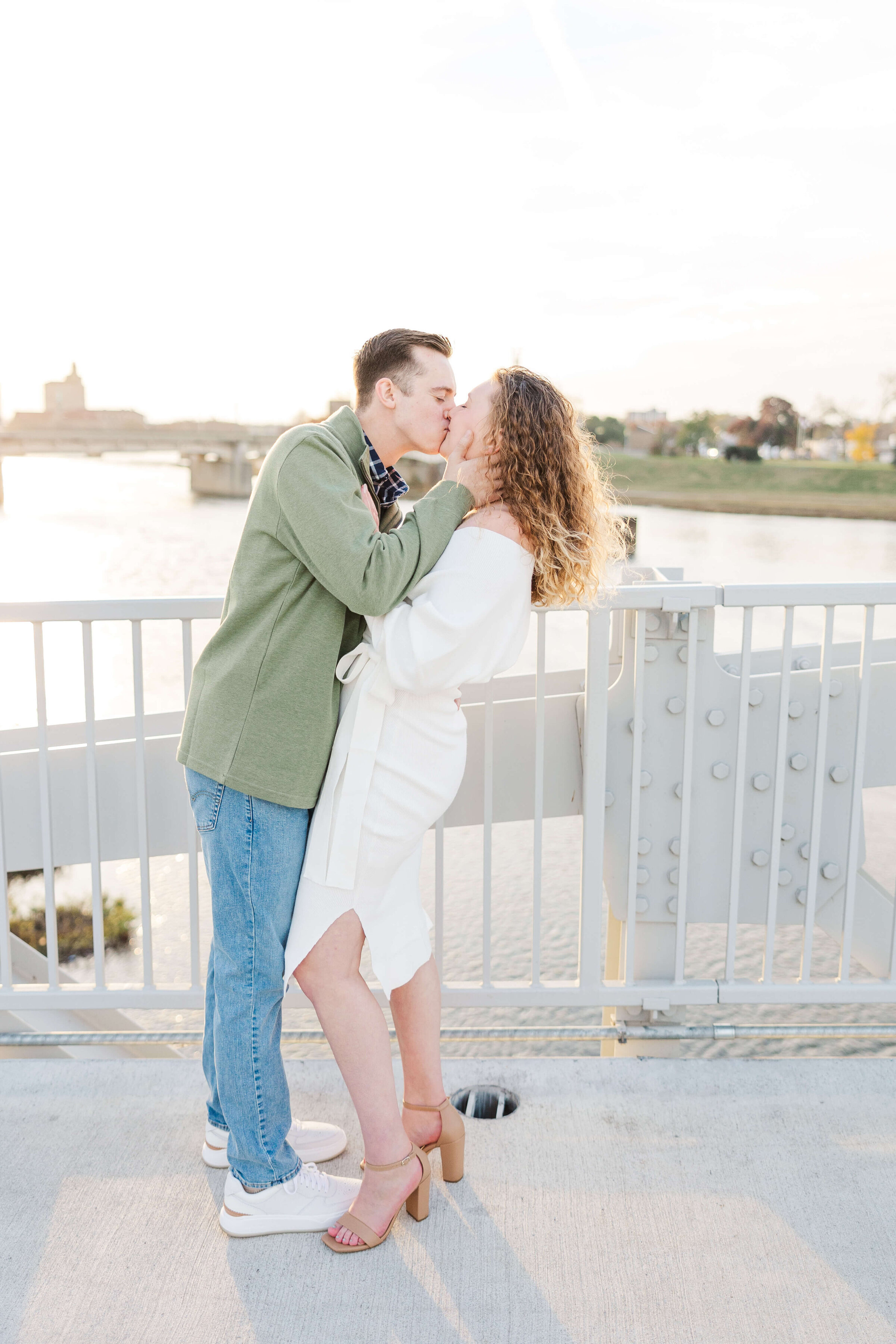 A man in a green sweater and jeans kisses his fiancee and dips her back. They are standing on a bridge in downtown dayton. Photo taken by a dayton wedding photographer.