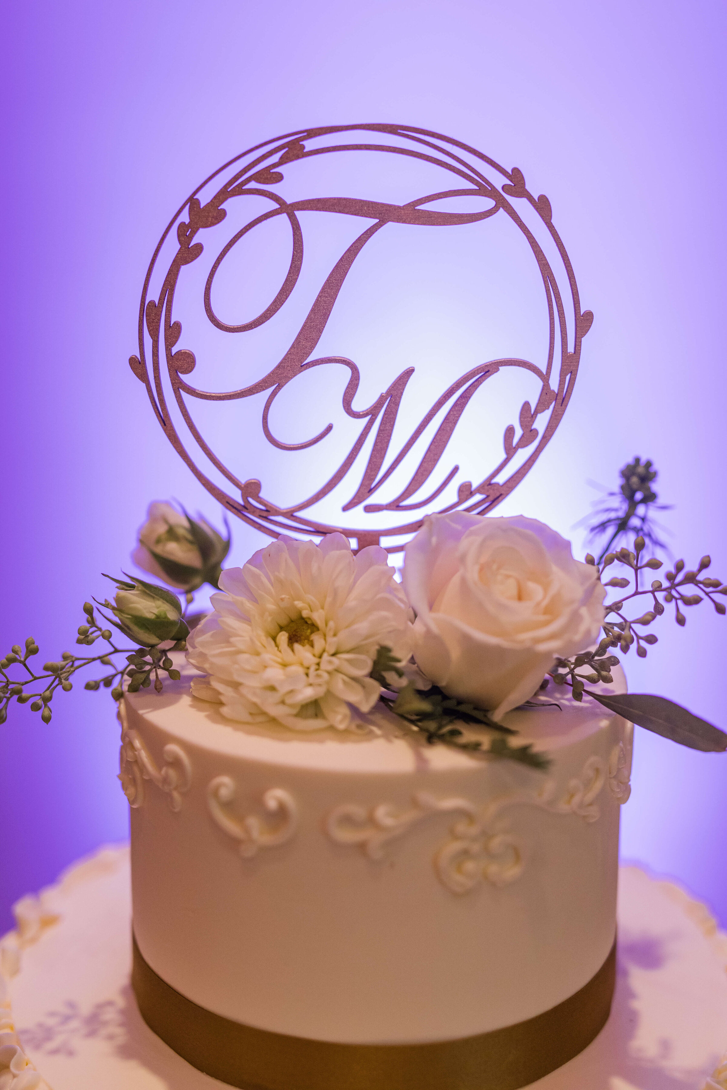 A wedding cake with a cake topper that has the initials T and M as the cake topper. There is a purple light behind it
