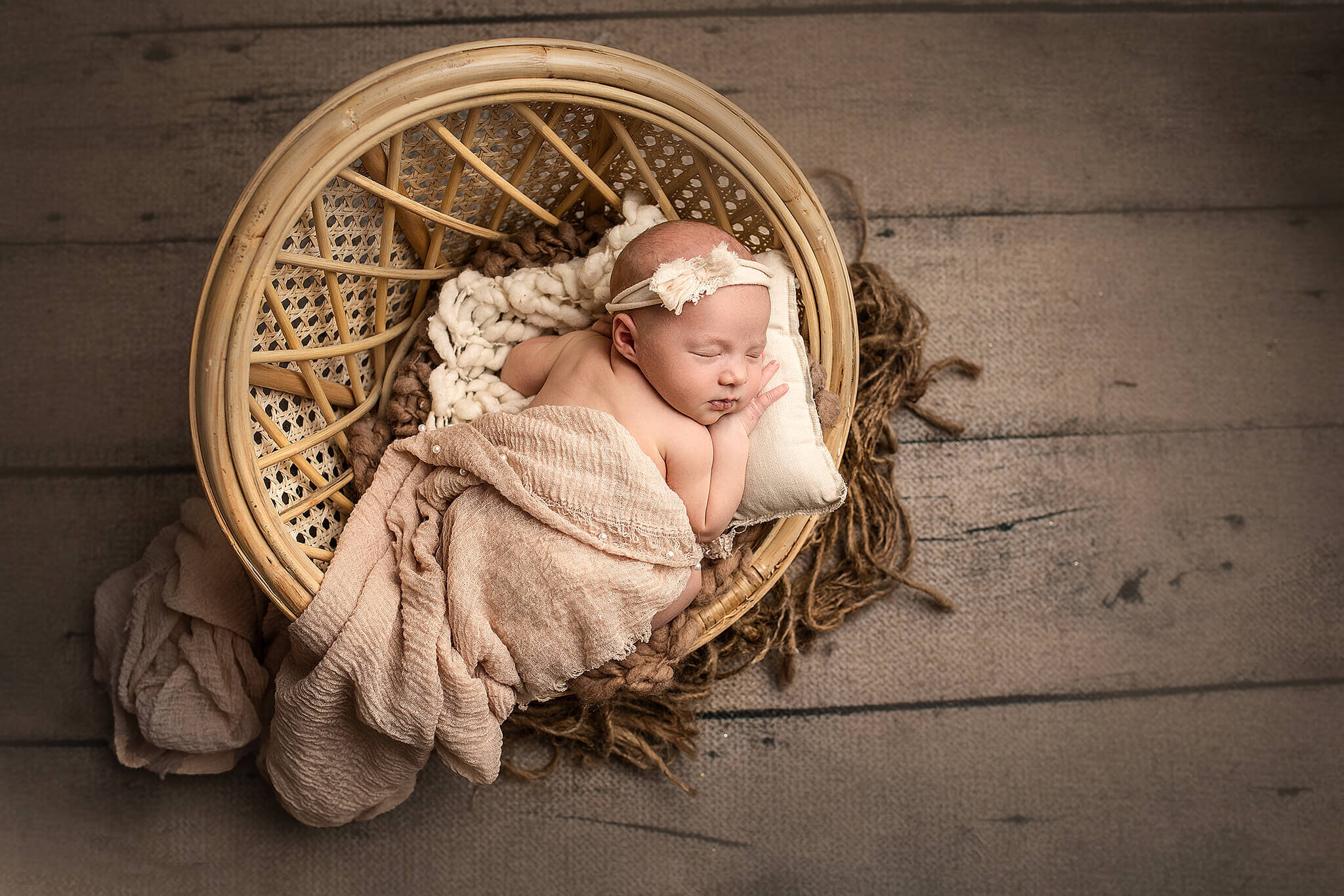 newborn baby girl sweetly laying in a round whicker chair while she is wrapped in a brown wrap, and laying her head on a little pillow