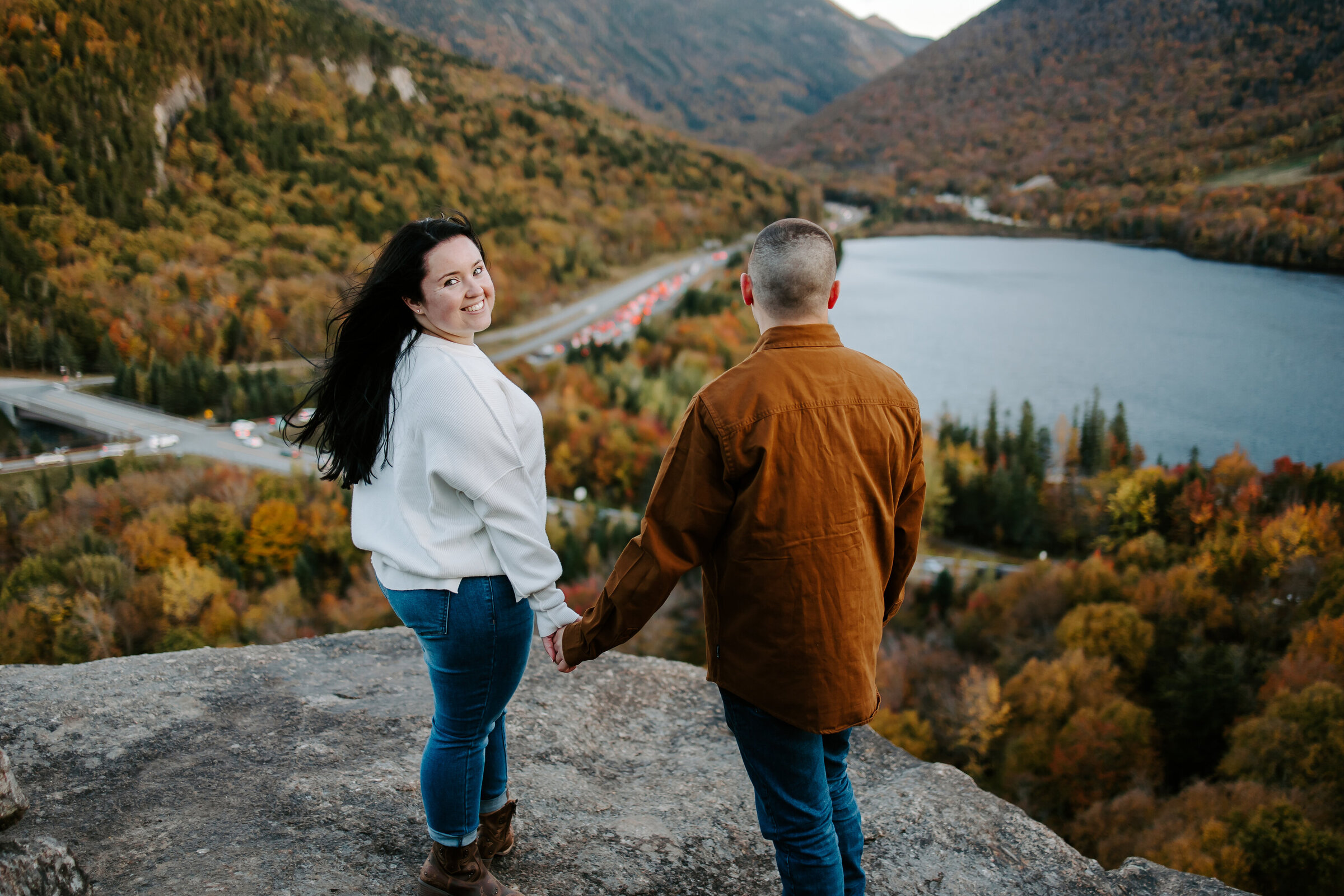 Bride to be looks over her shoulder at camera while holding fiances hand at the top of Franconia Notch overlooking Artists Bluff in lincoln, NH