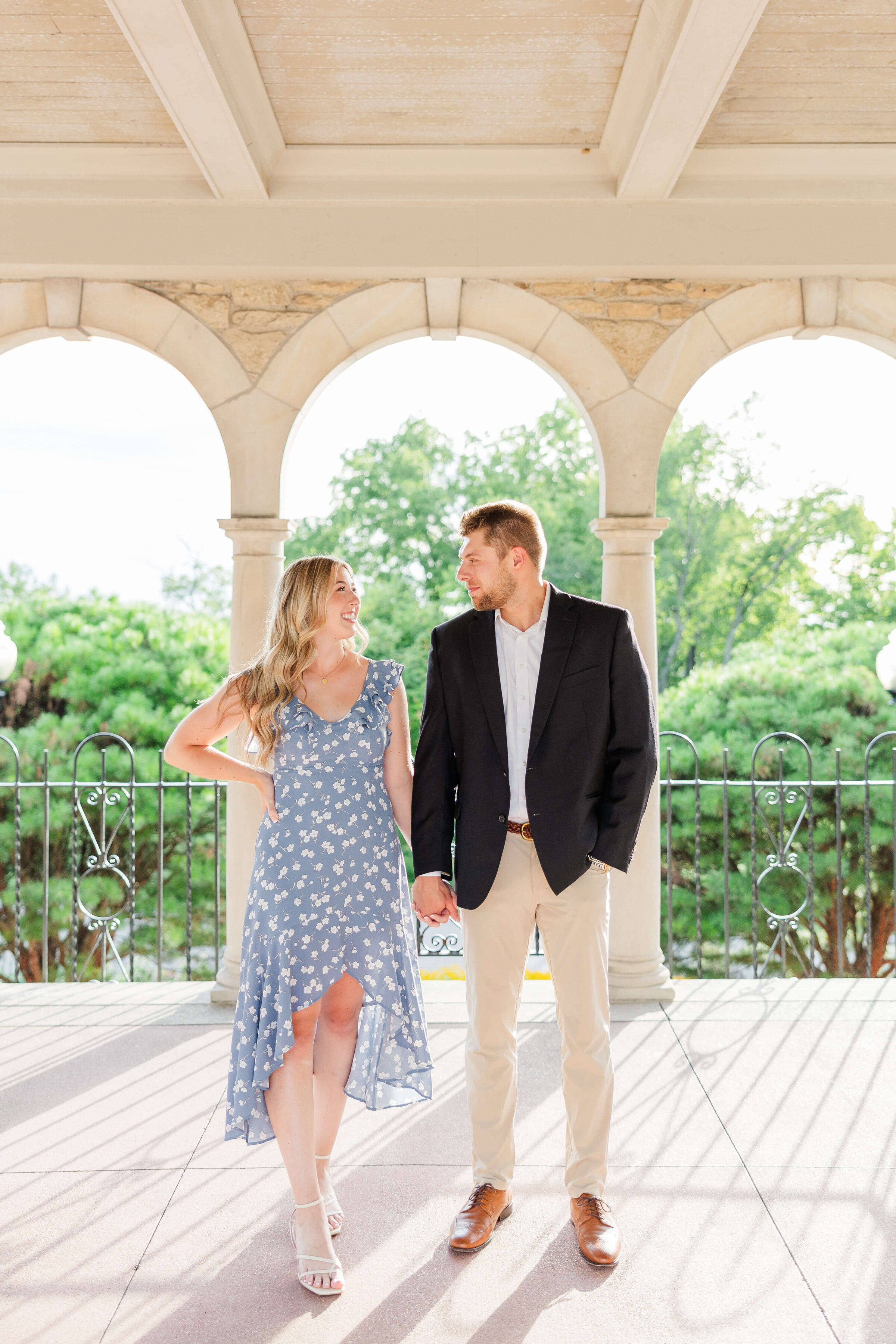 A woman in a light blue dress and a man in light pants and a dark jacket stand next to each other holding hands and smiling at each other. They are standing in a white building where the sun is glowing in and there are green trees in back of them. Taken by a cincinnati wedding photographer.