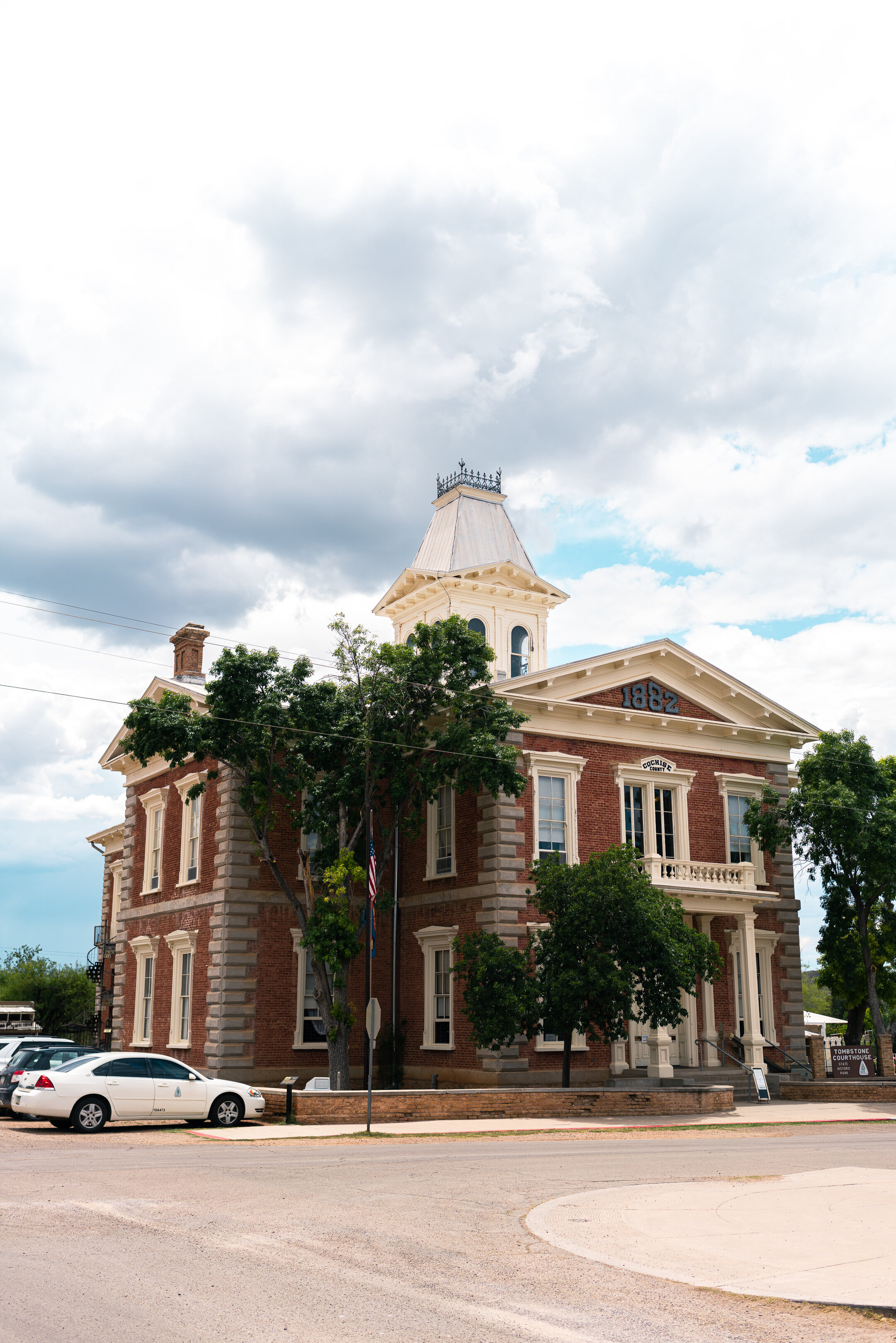 A red brick building with white trim and a tower, surrounded by trees and a road with cars.