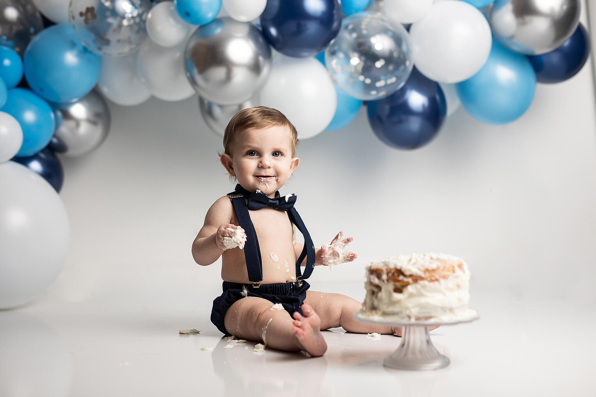 One year old little boy sitting on white flooring with white cake, and blue, silver and white balloons