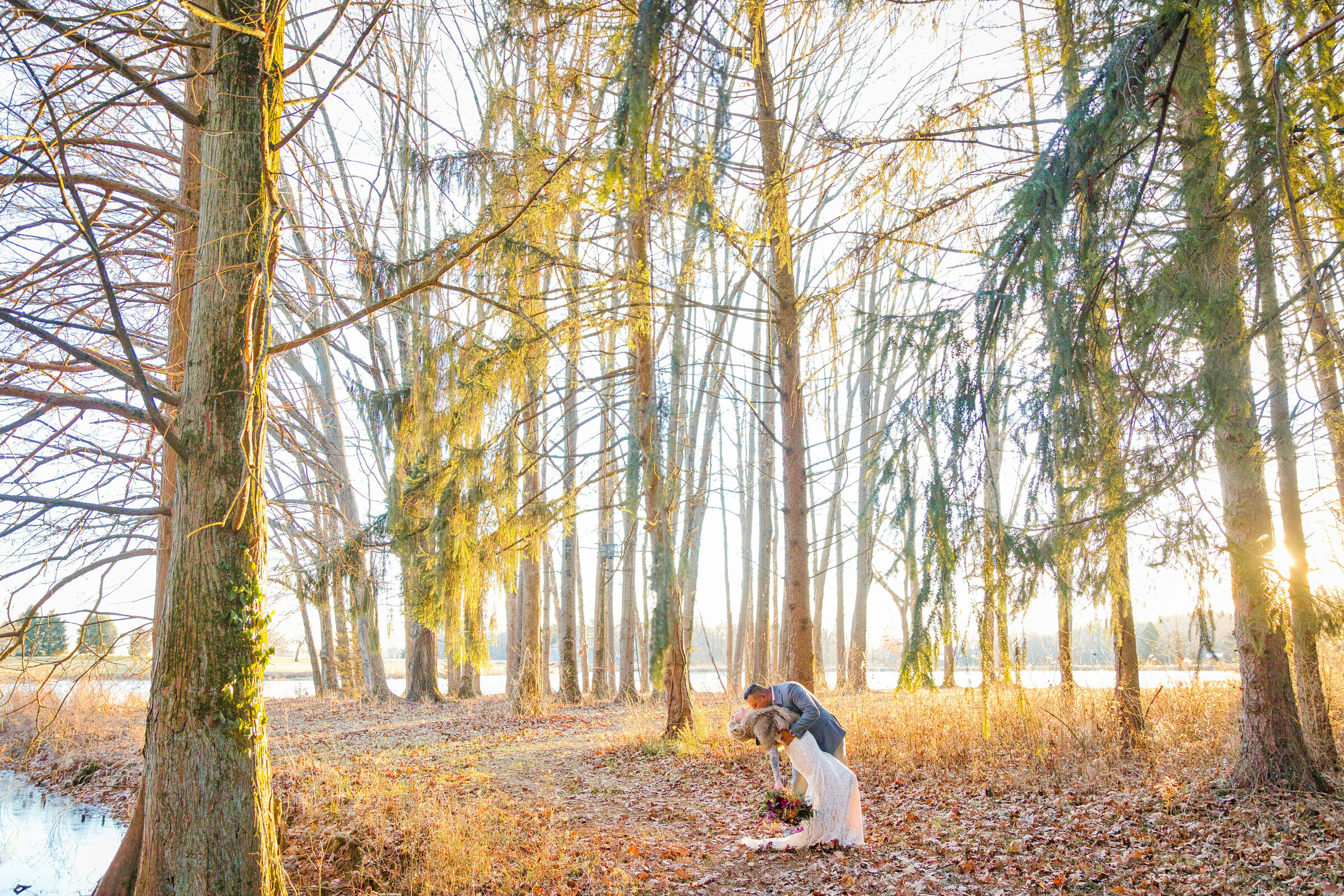 A bride and groom hold each other in the woods as the sun glows through the trees. He's wearing a grey suit and she's wearing a brown fur coat.