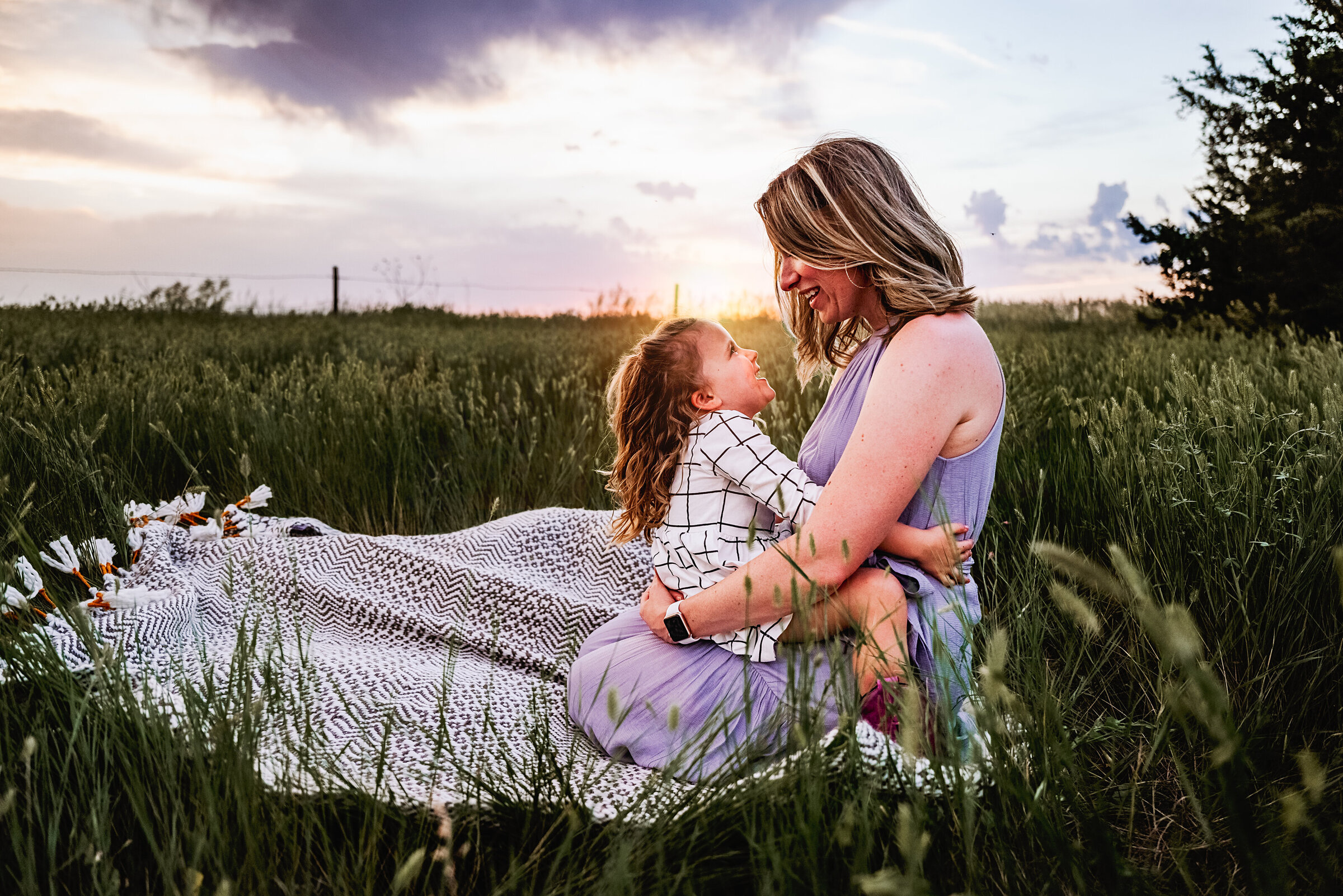 Mom and daughter sit on blanket in long grass smiling at each other