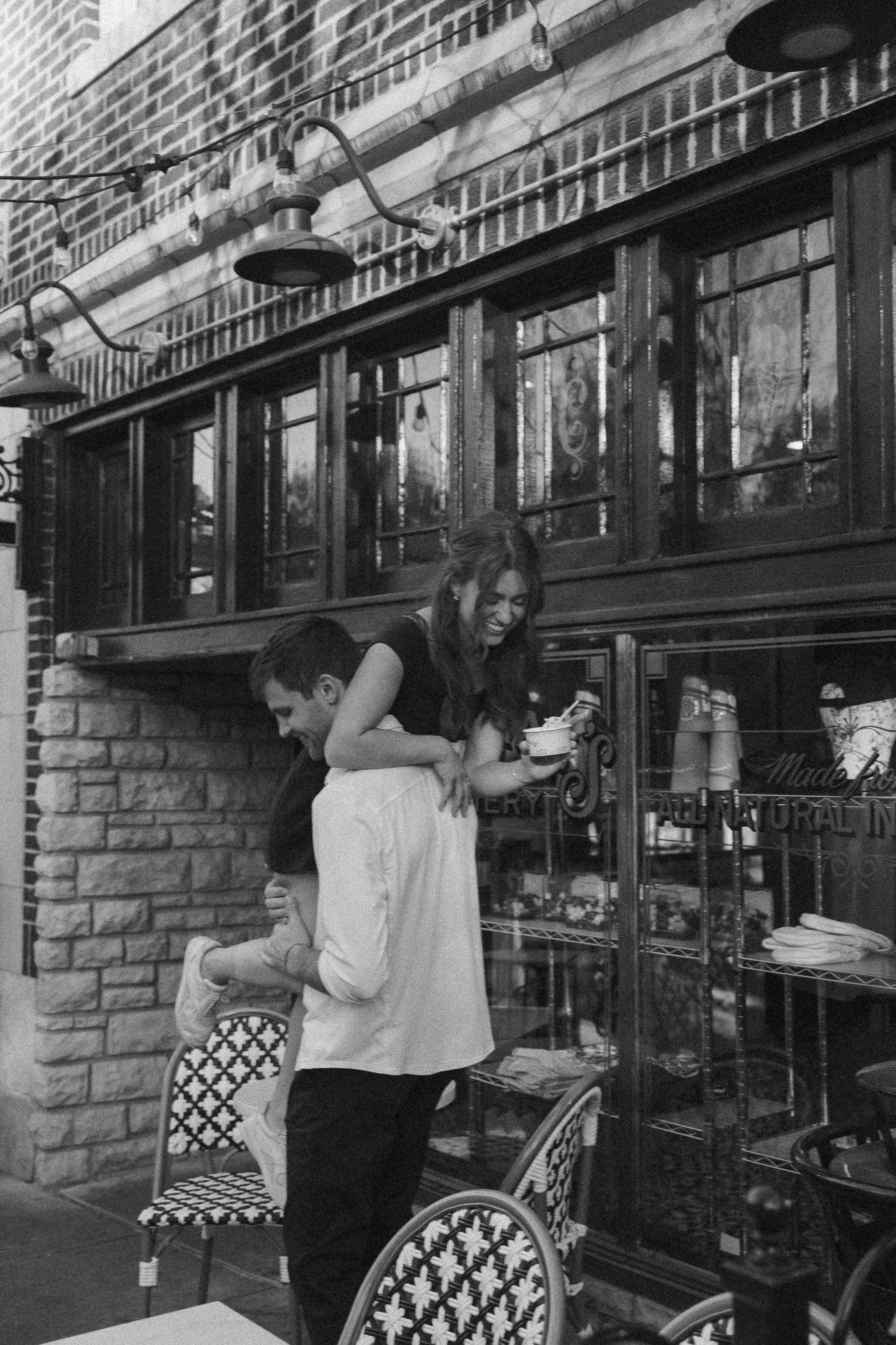 Monochrome shot of a man piggybacking a woman by a café.