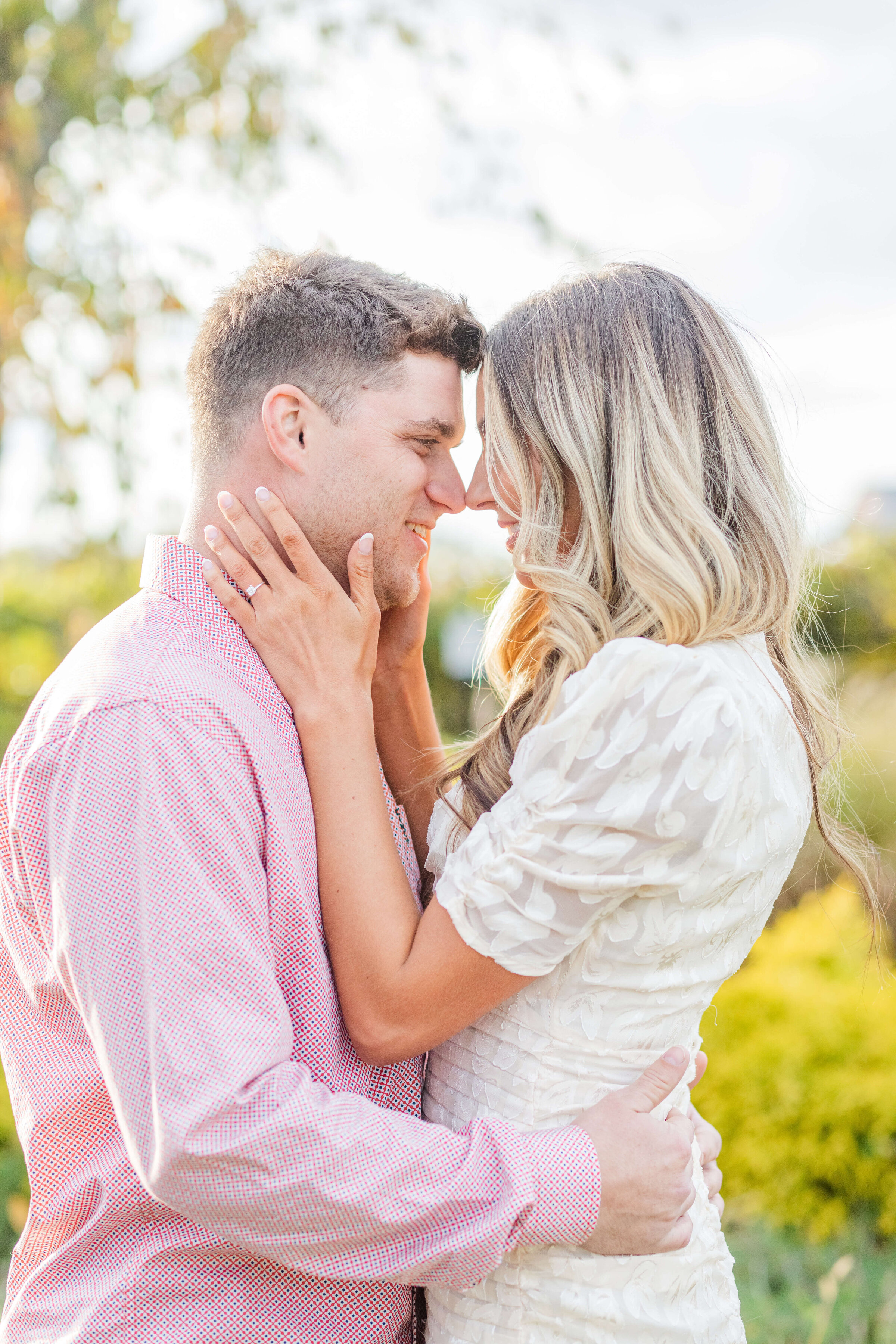 A man in a pink shirt and a woman in an ivory dress stand nose to nose as she has her hands on his face about to pull him in for a kiss. The sun is glowing behind them.