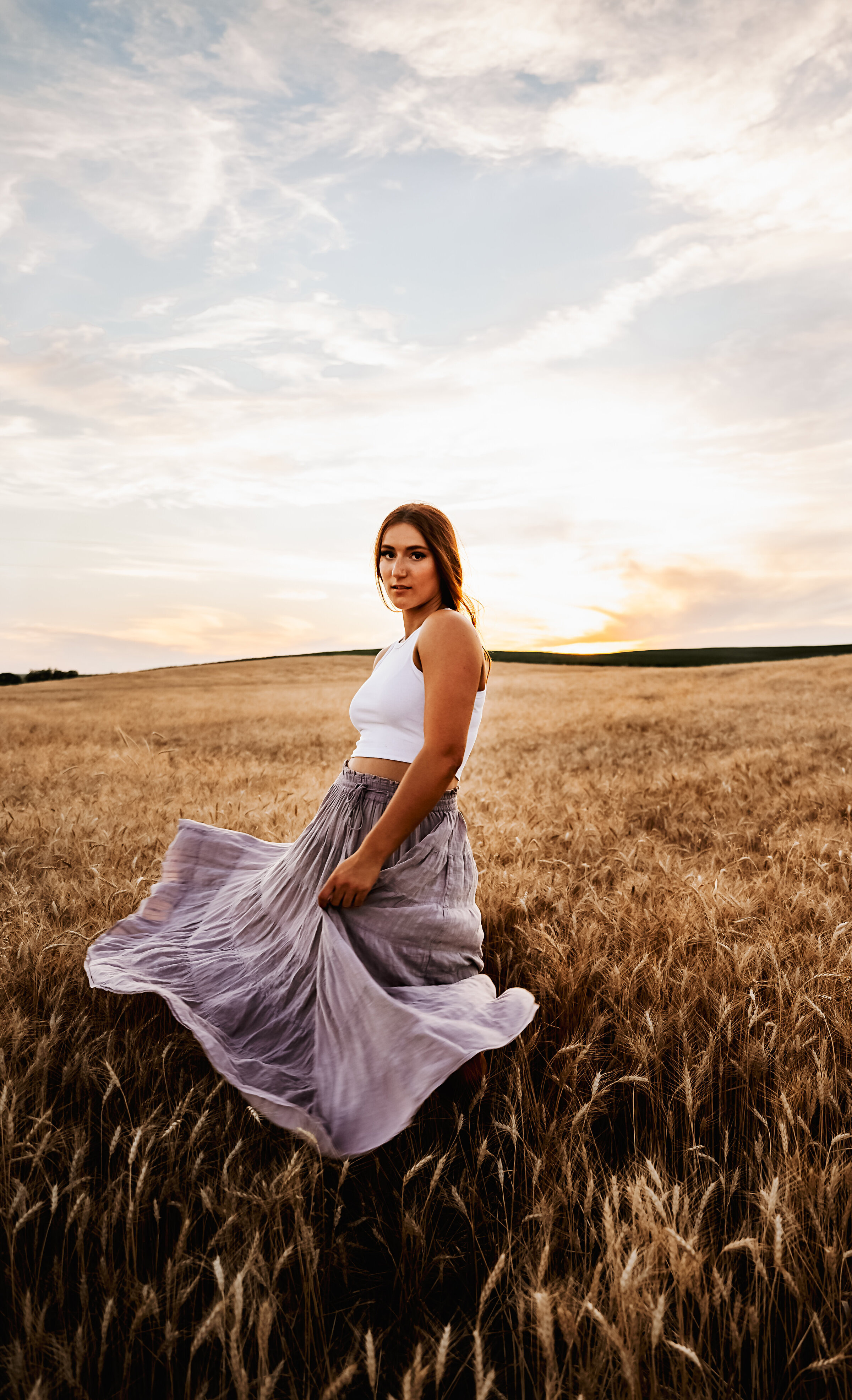 Girl twirls her skirt in golden wheat field with rolling hills at sunset