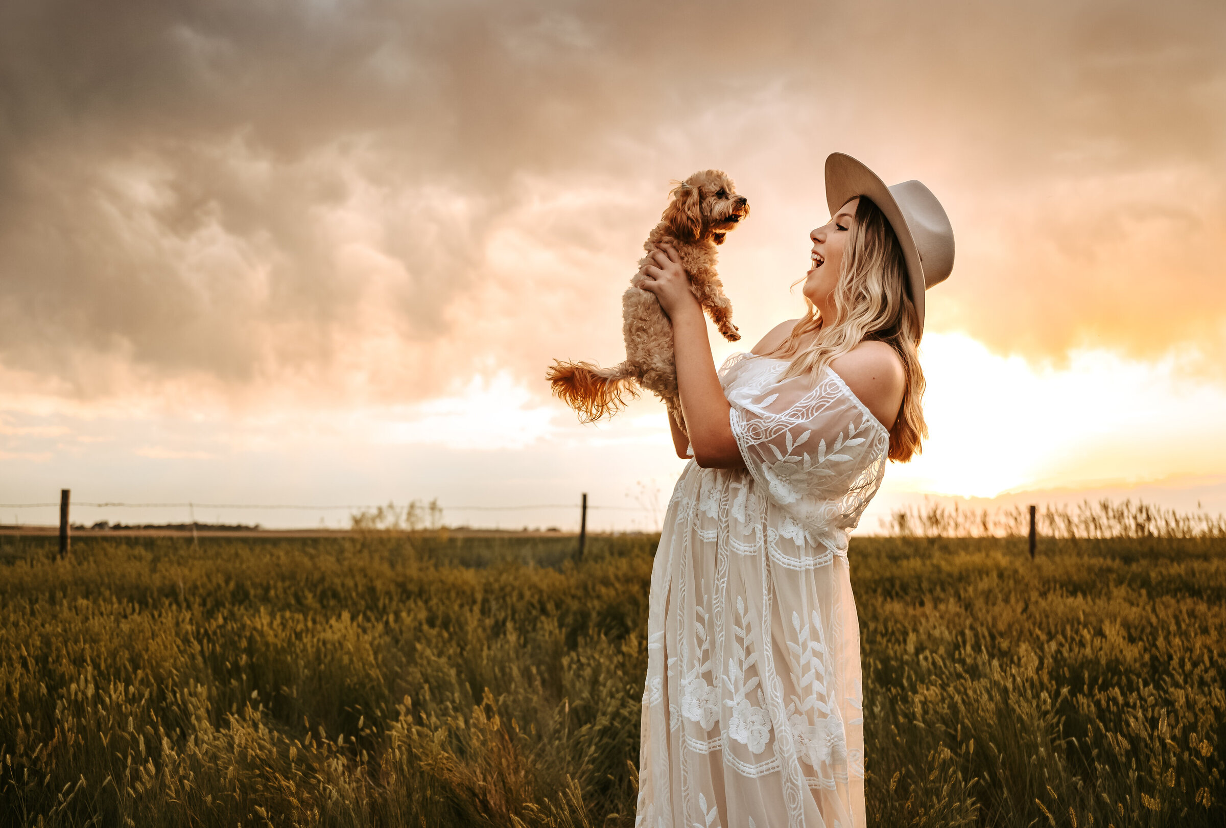 Senior girl in long white lace dress and wide brim hat hold little poodle dog up in the air and smiles at it