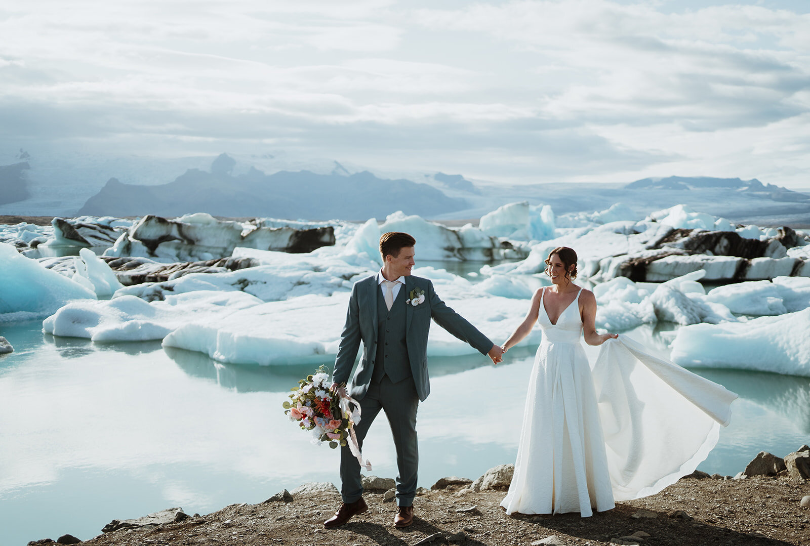 the bride and groom are walking together holding hands and smiling. the background is glaciers and sunshine. the bride has her dress flipped up and the groom is smiling at her while holding her flowers.