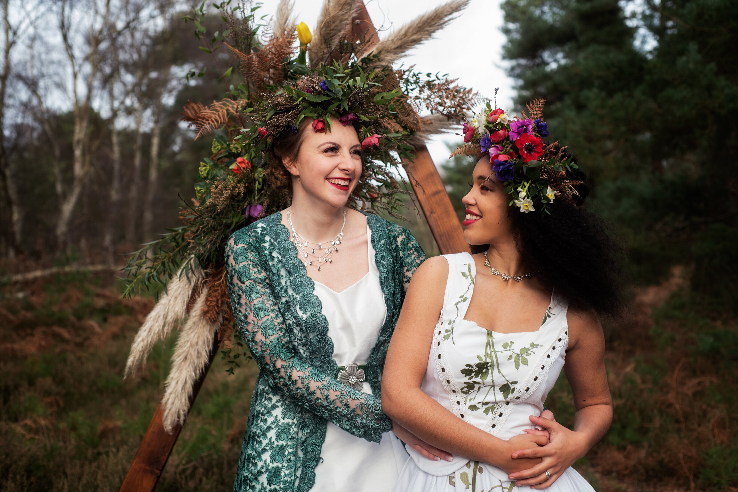 A pair of brides, clad in unique handmade upcycled wedding gowns, share a playful and loving glance in a whimsical forest setting, stood in front of their wedding arch adorned with vibrant and intricate florals. Both wearing floral crowns as they celebrate their elopement surrounded by the natural beauty of the woods.