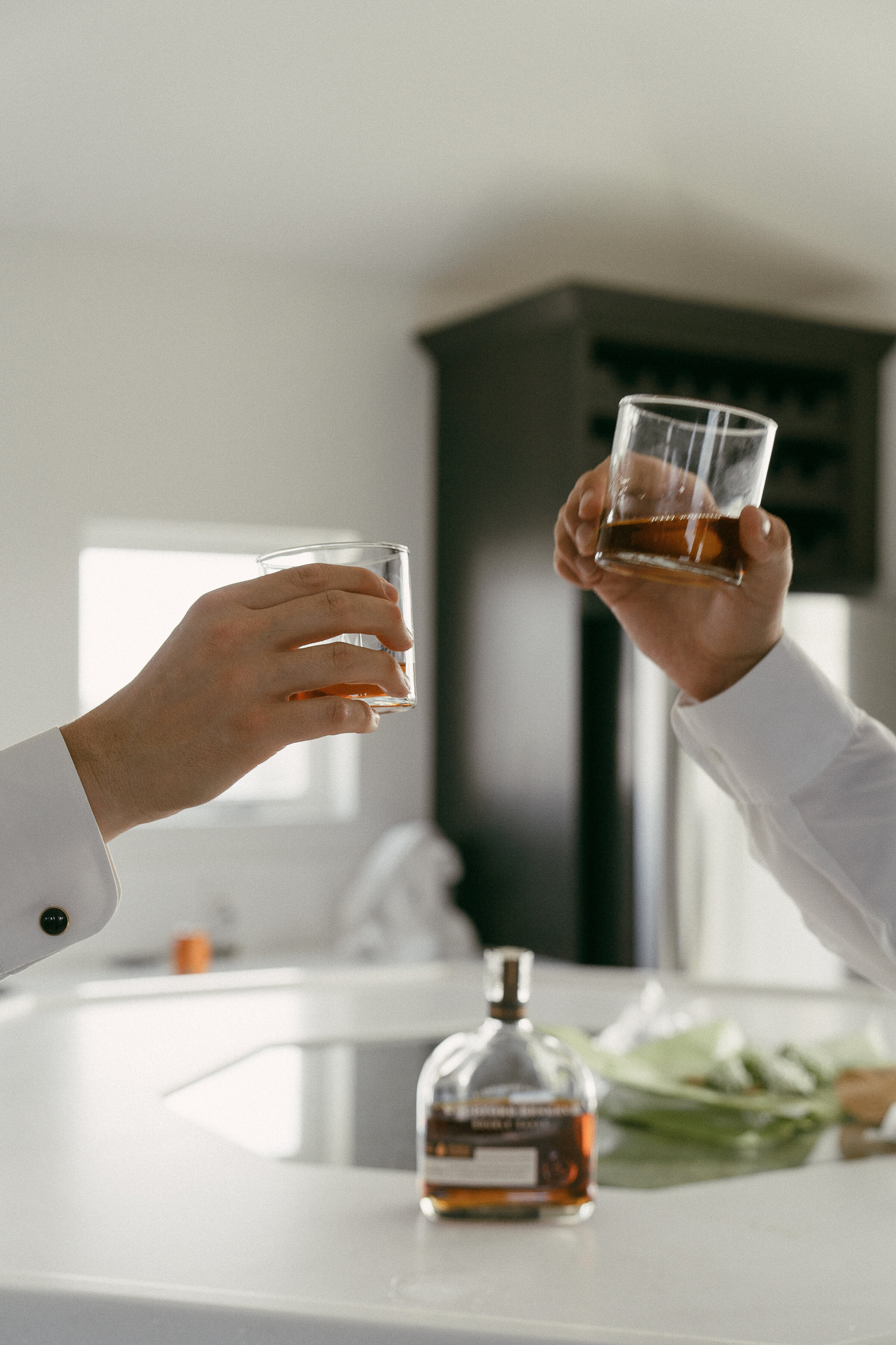 Close-up of two hands toasting with whiskey glasses above a kitchen island.