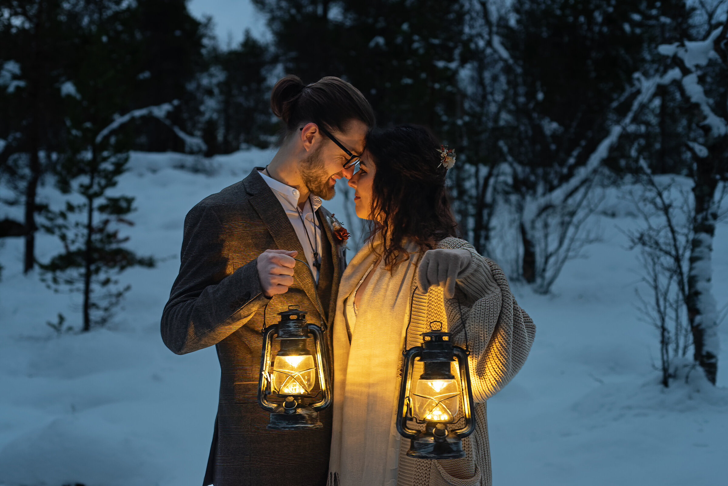 A romantic image of a couple who have just eloped in the Tromsø region, standing facing each other with foreheads together, in a snowy landscape during twilight. The man, wearing glasses and a stylish brown tweed suit, is holding a lit lantern in his right hand. The woman, adorned with small flowers in her hair and a cozy cream cardigan over a white boho wedding dress, is holding a similar lantern in her left hand. They are gazing affectionately into each other's eyes, sharing a warm moment in the cold environment.