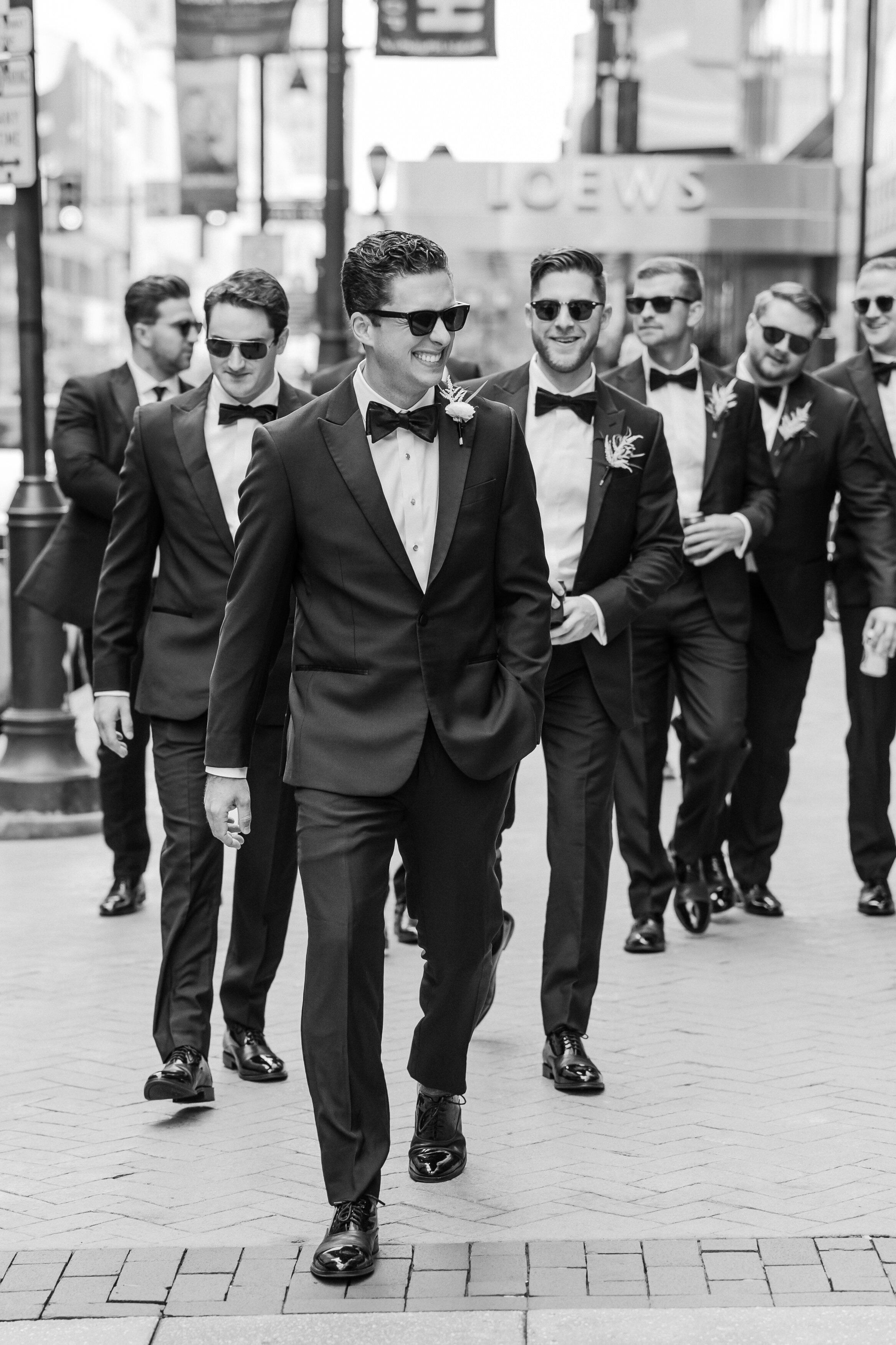 Groom along with his groomsmen in black tuxes and black sunglasses are walking down the street from the Loews Hotel in Philadelphia
