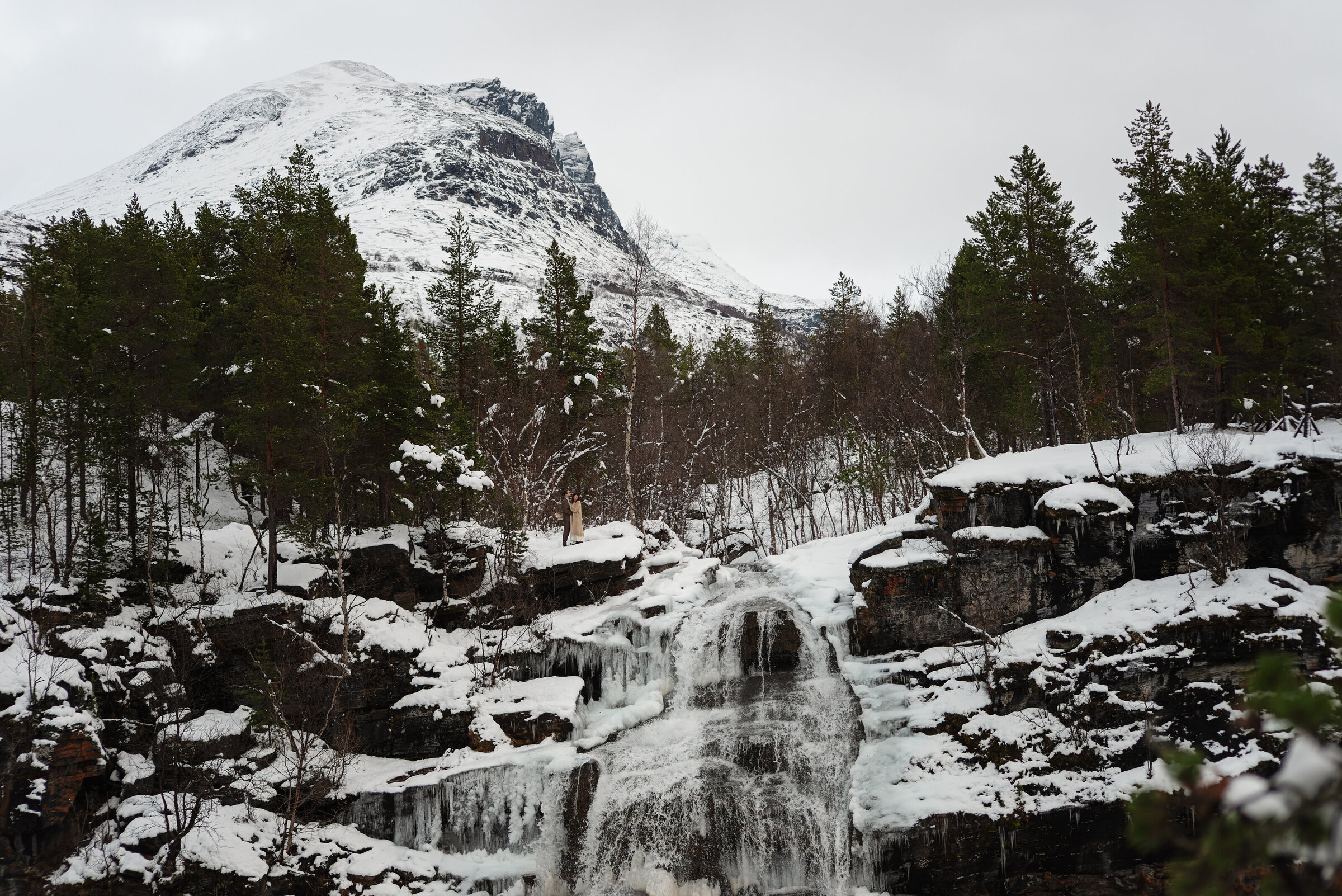 In the serene landscape of Norway, a couple stands at the top of a partially frozen waterfall. The grandeur of the mountain looms in the background, draped in snow, complementing the intimate moment that marks their elopement, with the wild beauty of nature as their witness.