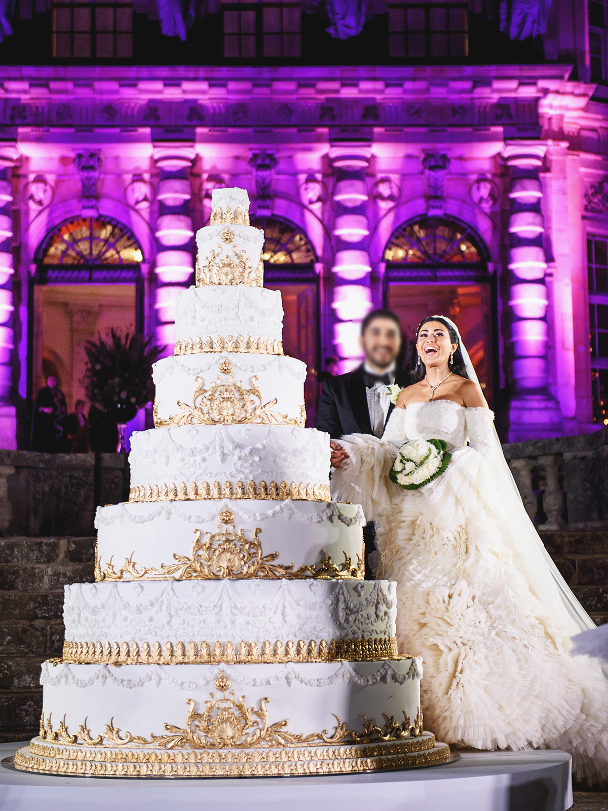 Bride and groom standing behind very tall white and gold wedding cake
