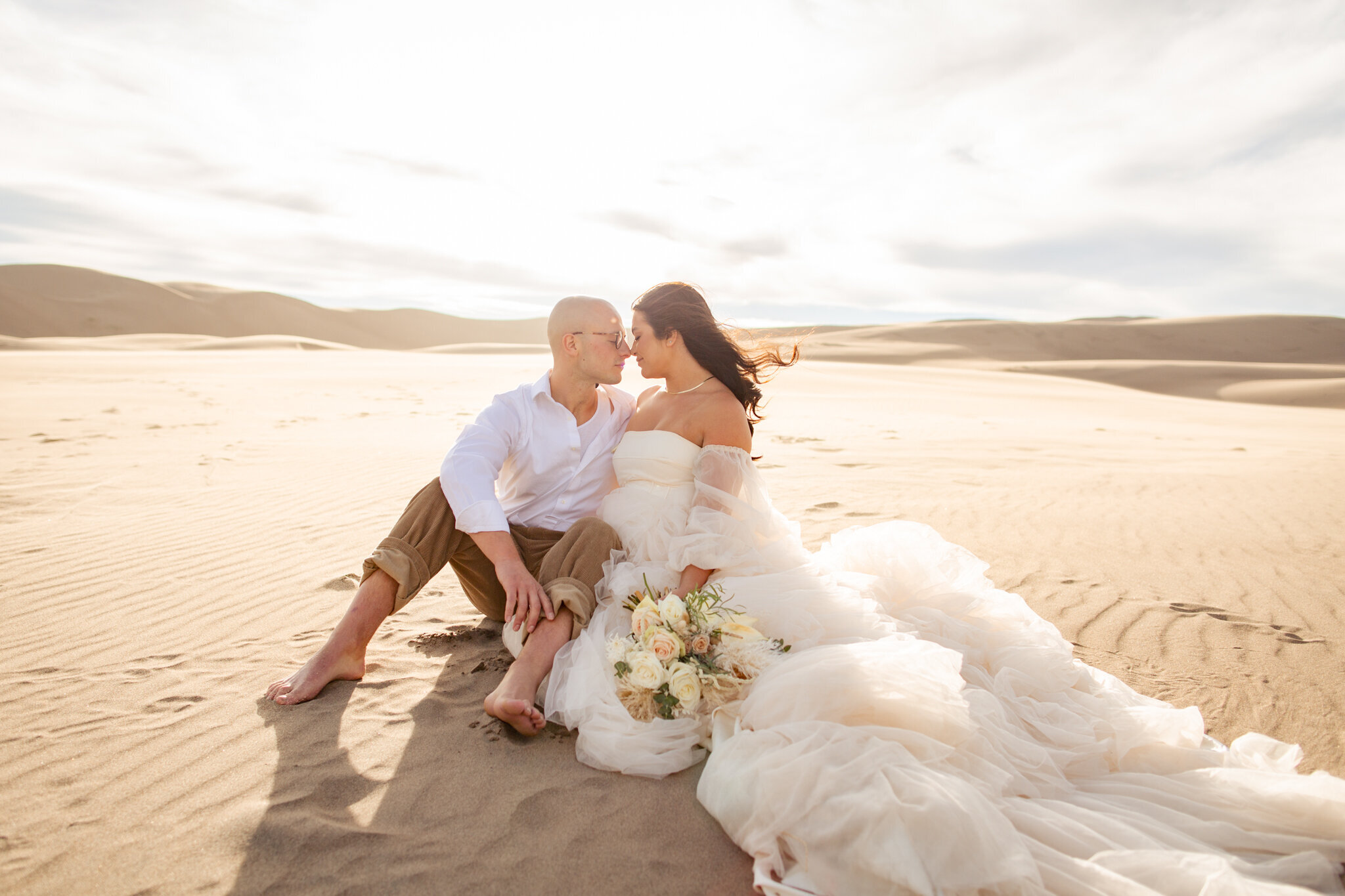 The bride and groom sit next to each other on the sand at the sand dunes.