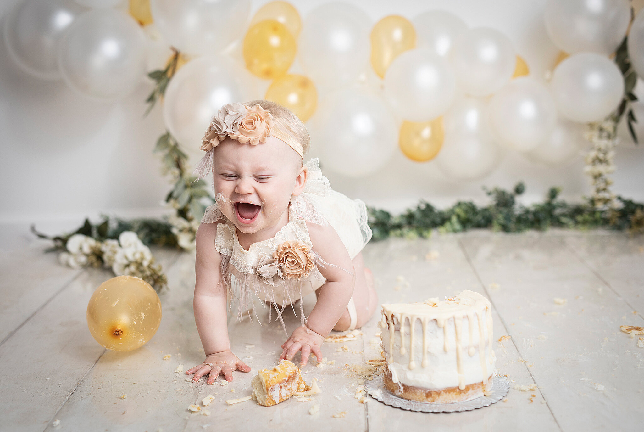 baby girl with white and gold balloon arch cake smash while baby is laughing