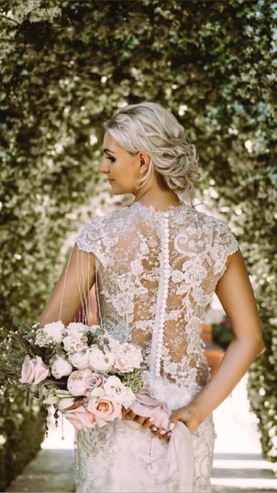 The back of a blonde haired woman holding a  bouquet of pink roses stands in front of two rows of trees.