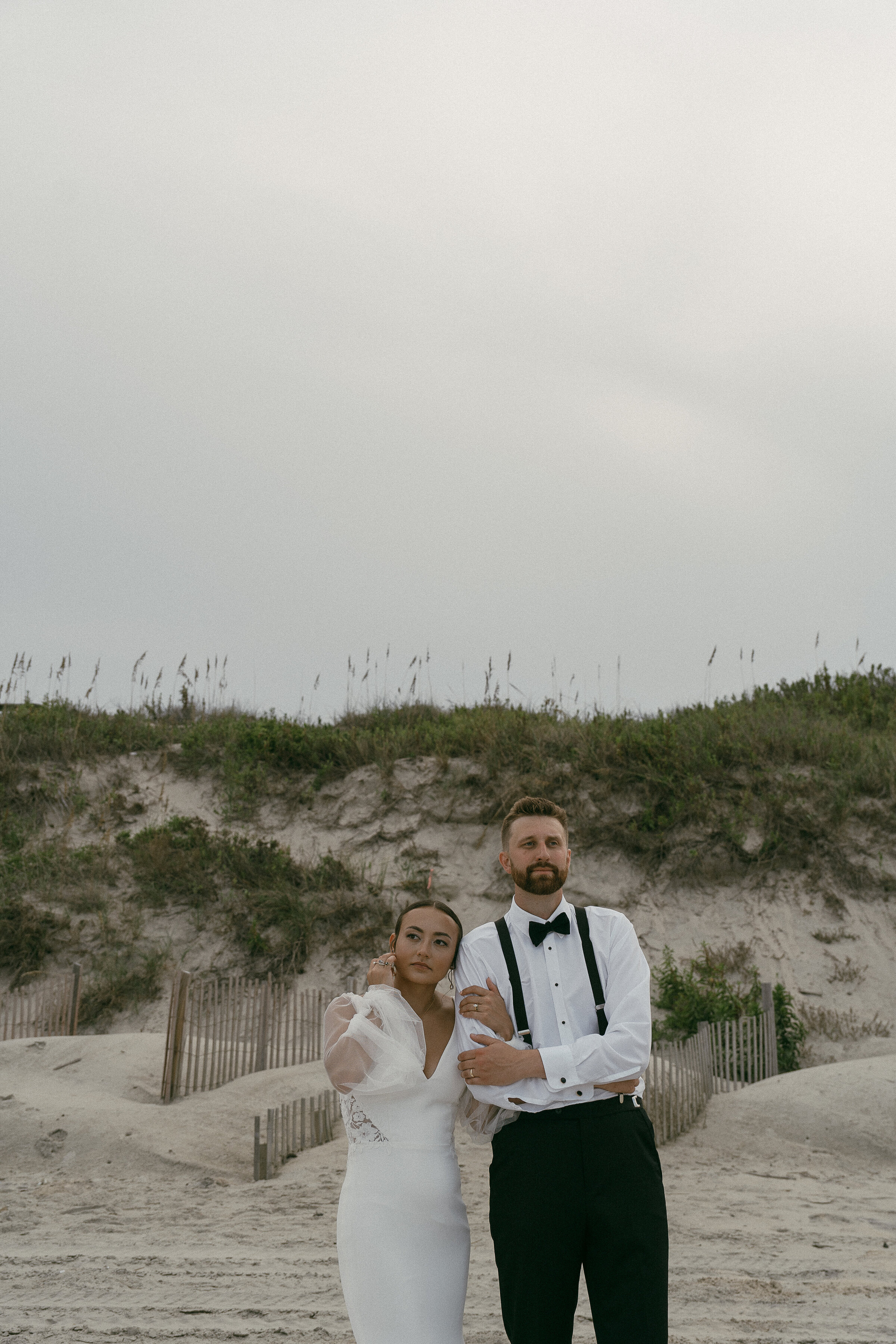 Bride and groom standing solemnly on the beach, with dunes in the background.