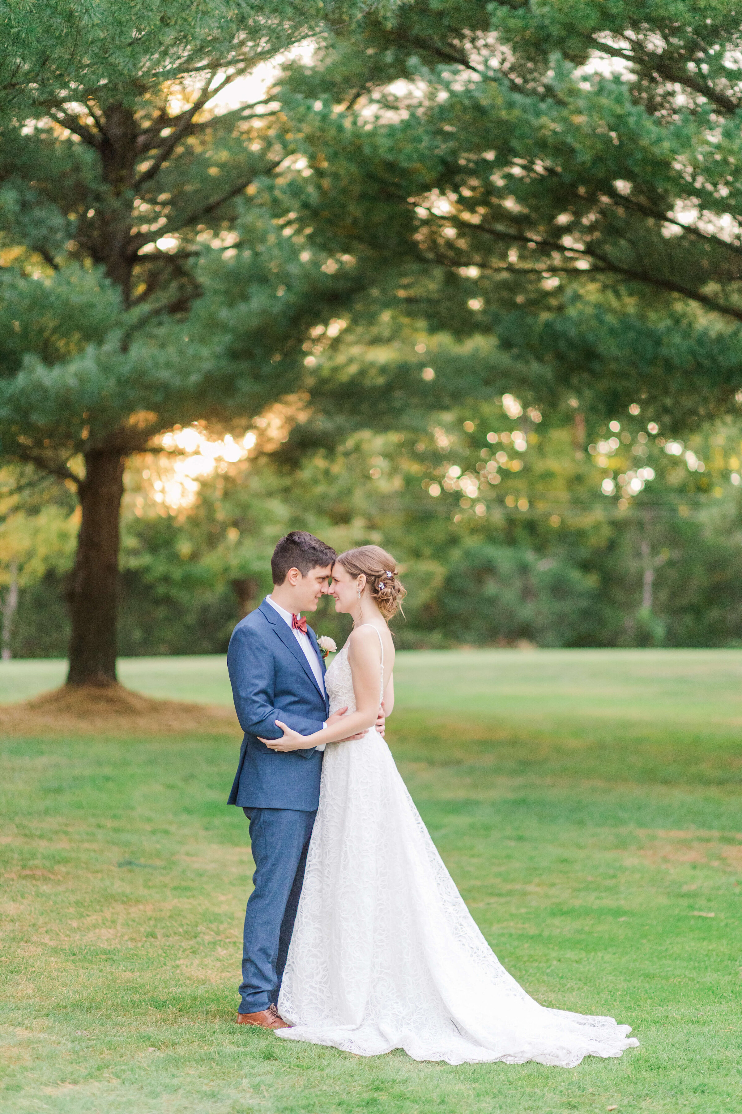 A bride and groom hold each other standing with their foreheads touching. They're standing in front of an evergreen tree as the sun glows through it.  He's wearing a blue suit.
