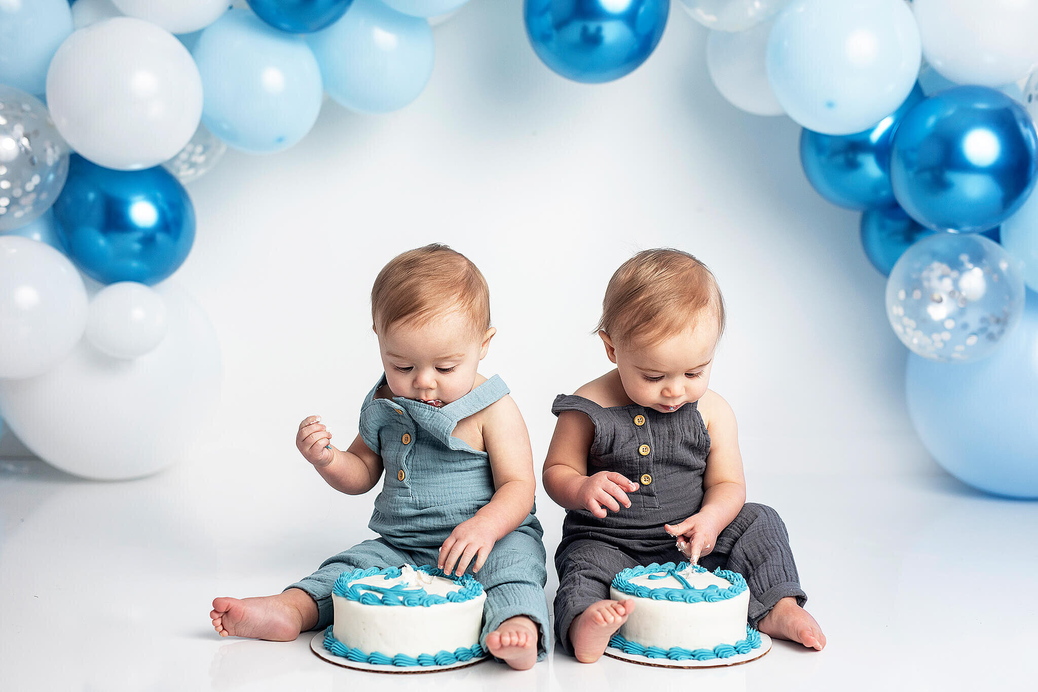 Twins dressed in blue overalls and white, silver and blue balloon arch on the background