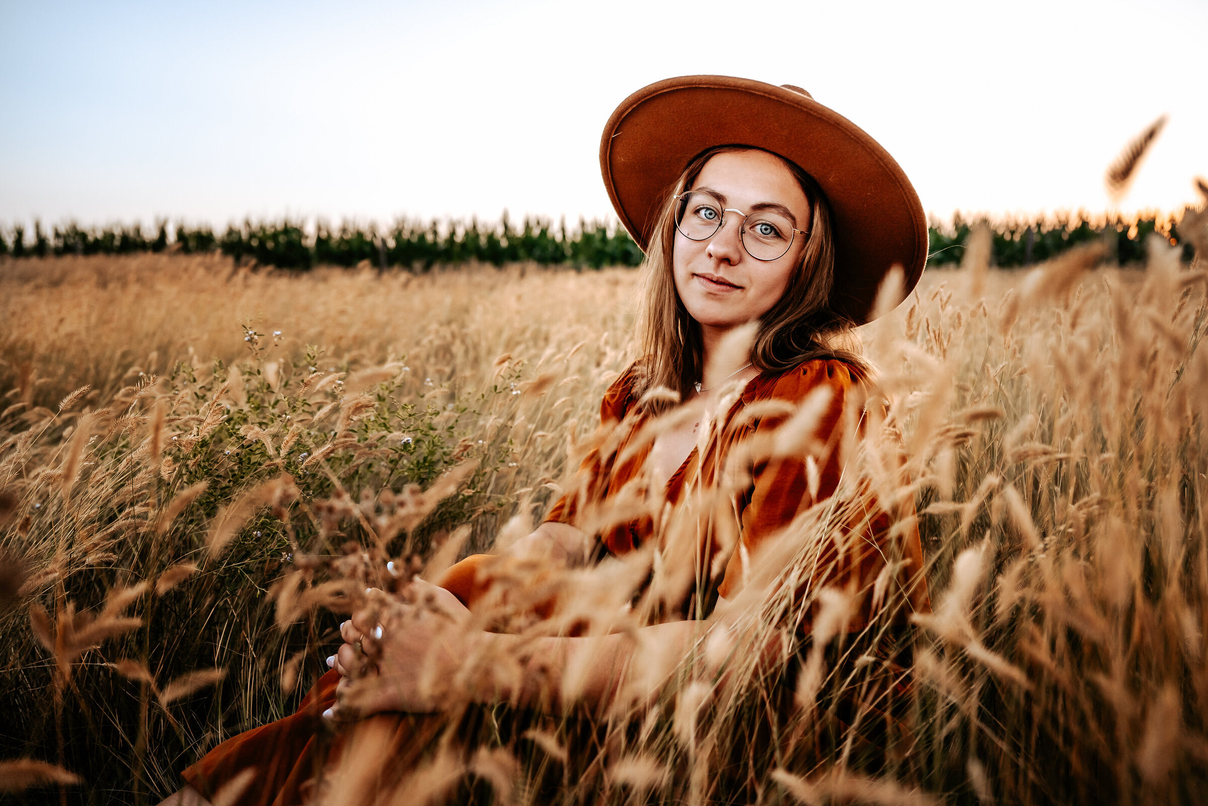 Girl with wide brim felt hat, round glasses, and rust colored dress sit in long grass
