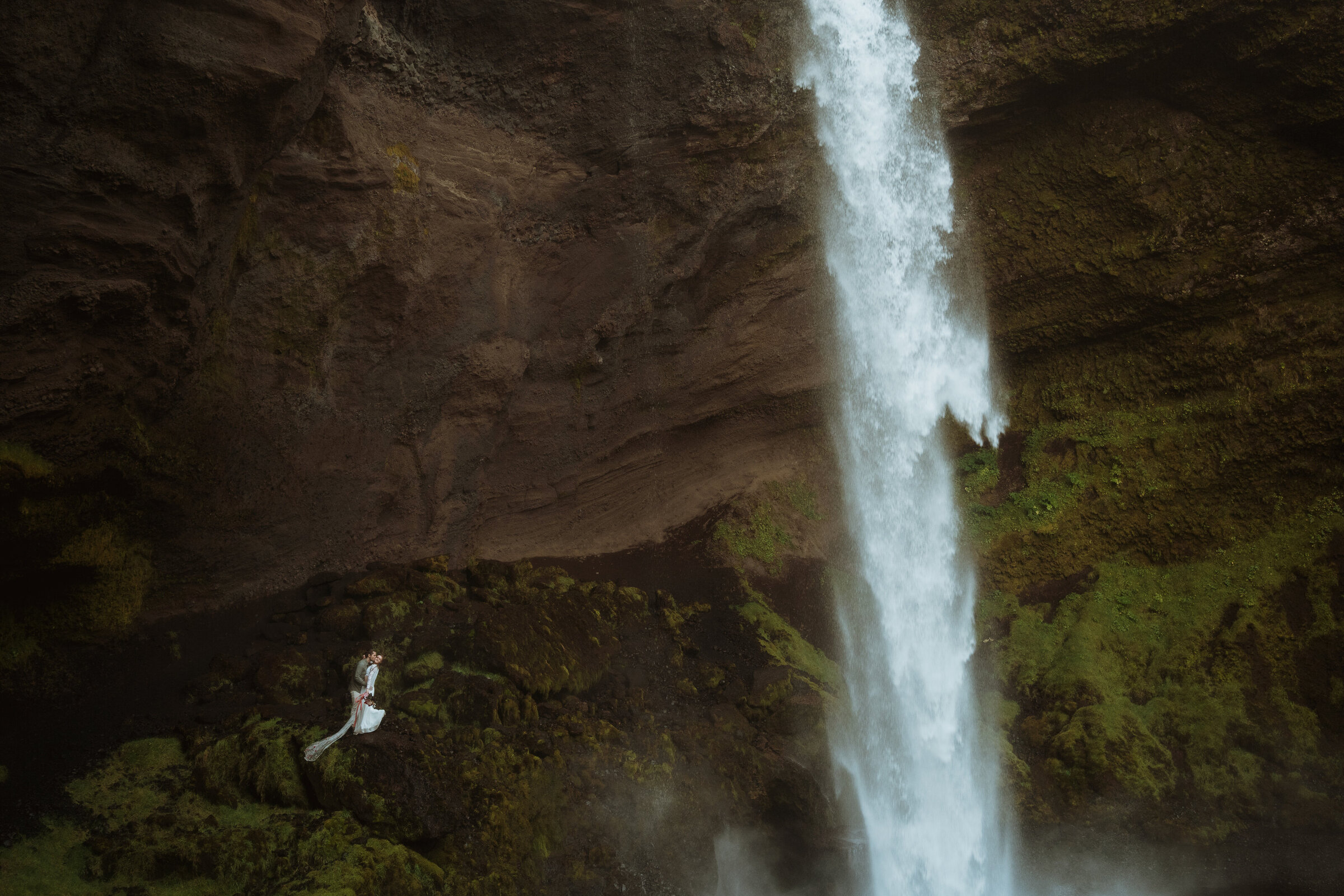 the couple is standing behind latourell falls in the columbia river gorge. the groom is dipping the bride and kissing her cheek. they are far away from the camera surrounded by rocks and greenery as the waterfall is to the right of them.