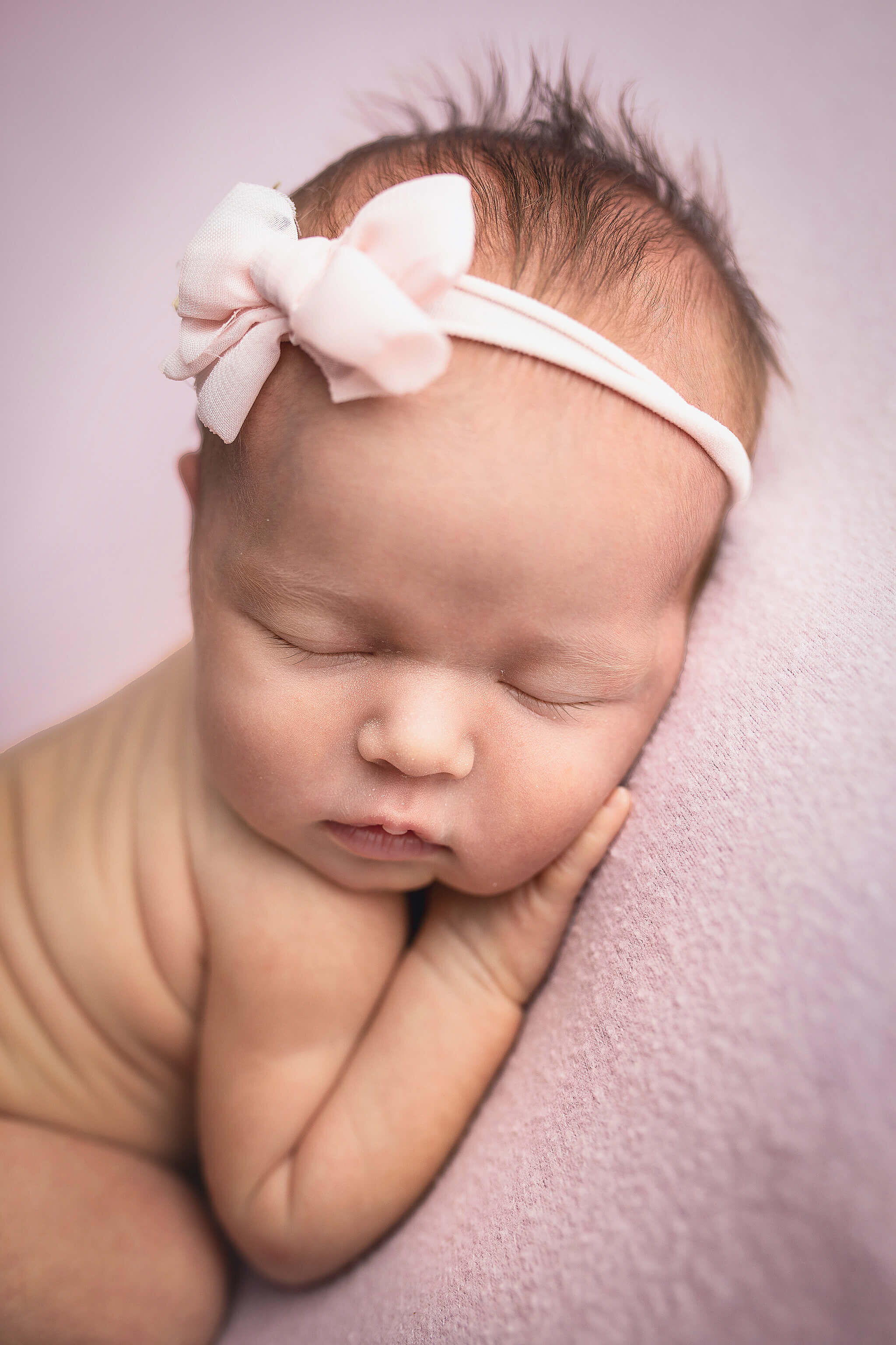 newborn baby girl laying on a pink blanket while sleeping.  pink bow on her head.