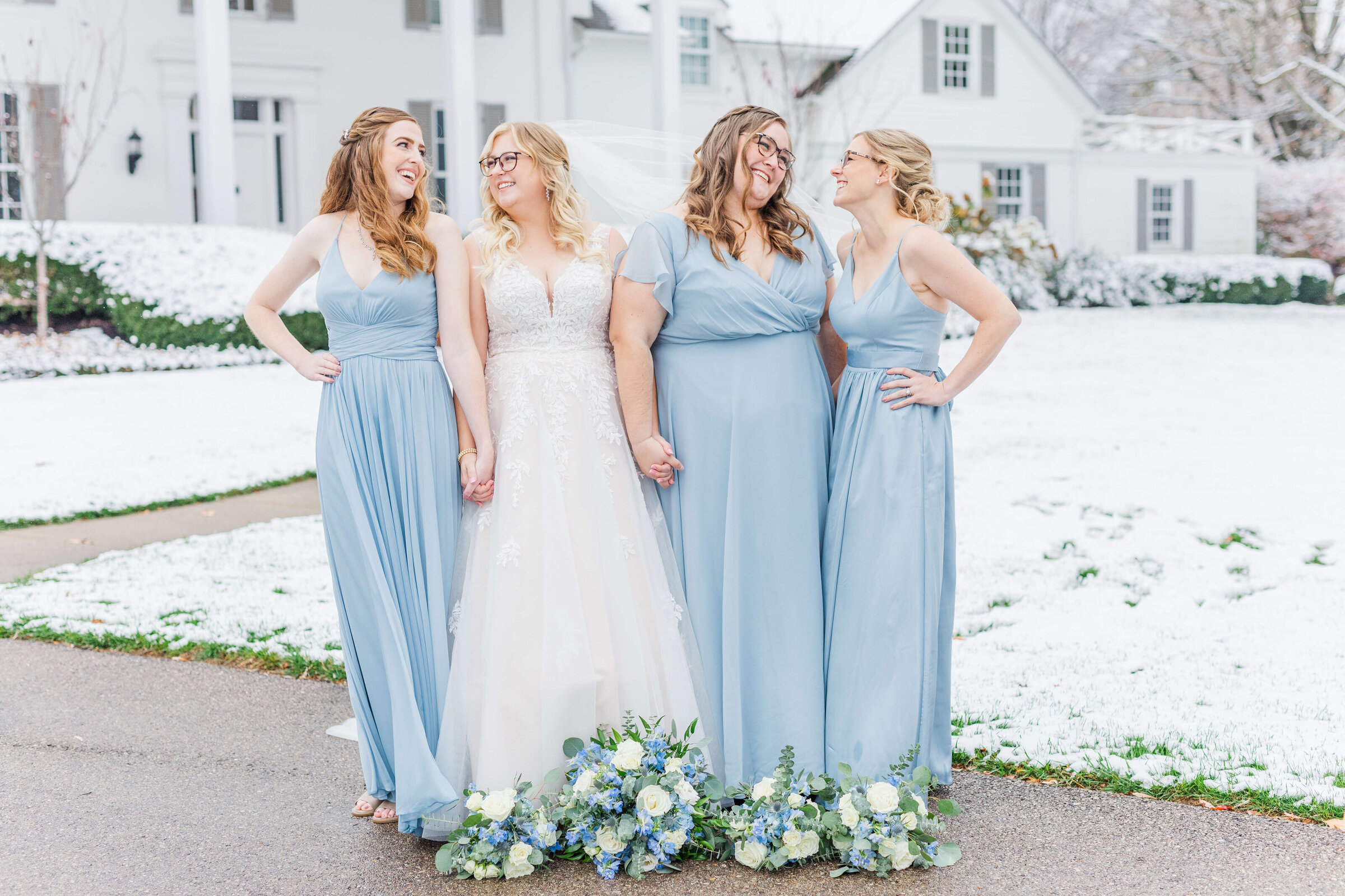 A bride and her bridesmaids in light blue hold hands. It just snowed so the entire background is white and snow covered. Taken by a dayton wedding photographer