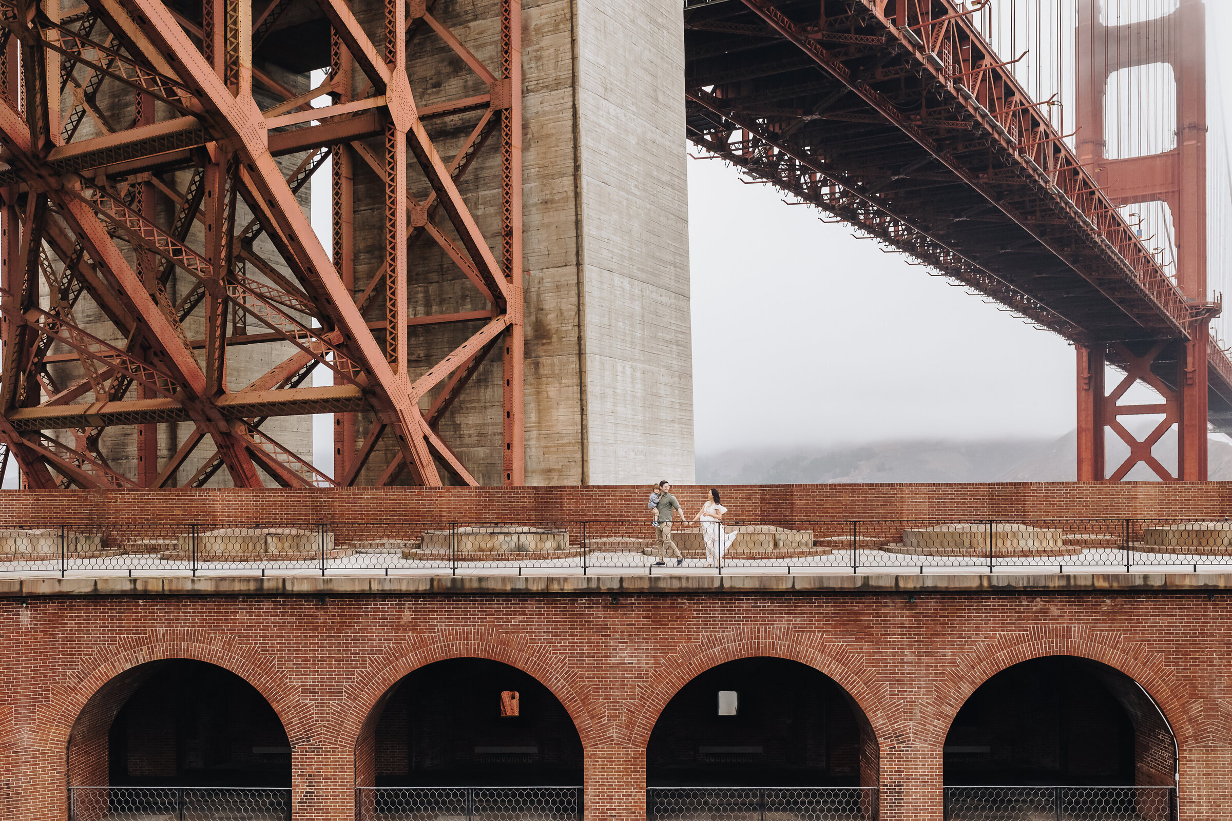 A family walks under the golden gate bridge during a maternity session at Fort Point, San Francisco