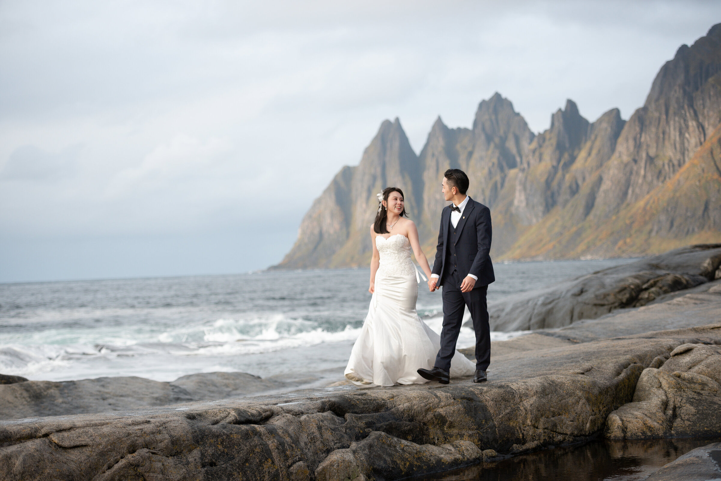 A newlywed couple holding hands and walking on the rocky shores of Senja, Norway, with towering, jagged mountains in the background and dramatic clouds above, celebrating their elopement in this rugged landscape.