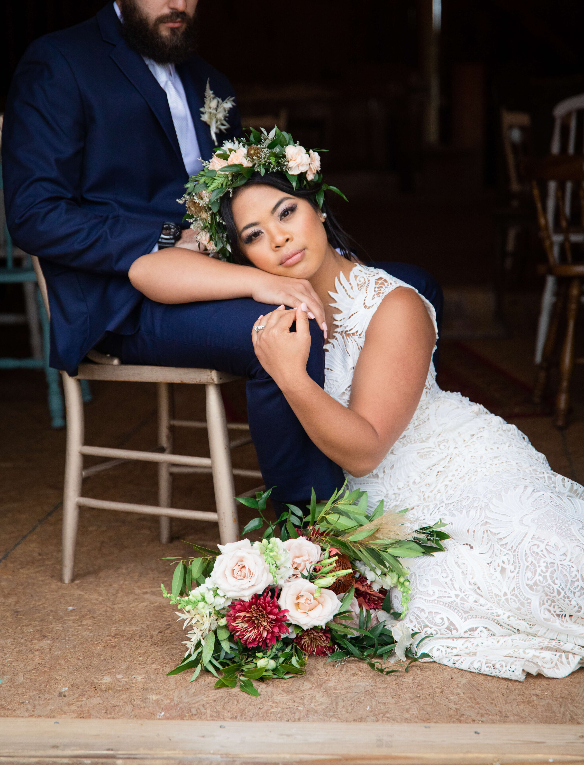 A bride is sitting on the floor while her groom sits on a chair next to her. She is resting her face on his leg and looks content and happy. She is wearing a white dress, a floral head crown, and a bouquet of flowers on the floor.