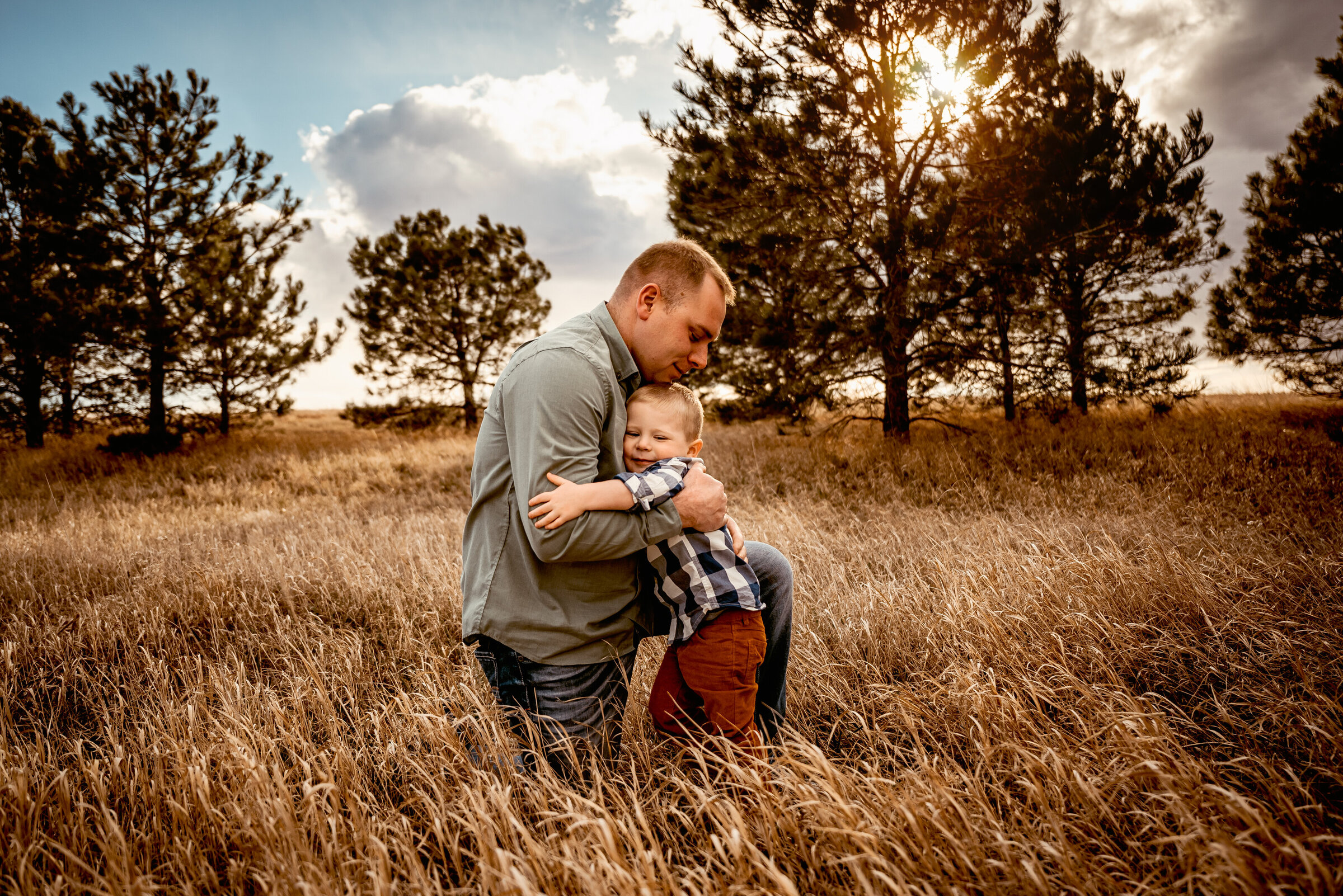 Dad and young son hug each other in field in front of pine trees