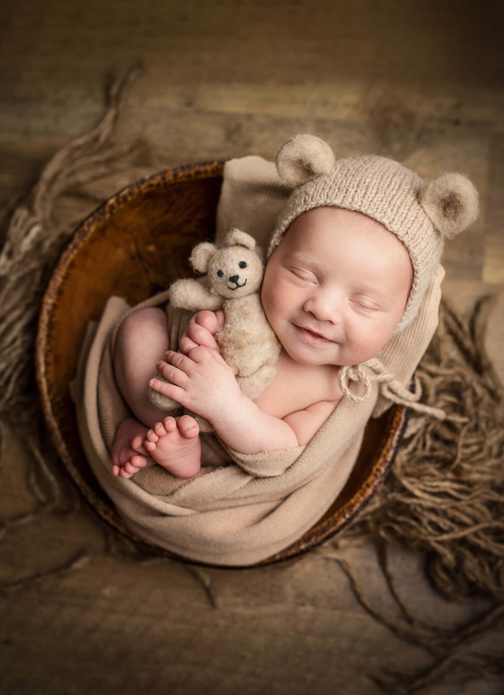 newborn baby boy wrapped in tan wrap, with a little bear hat on and snuggled with a little bear, while laying in a little bowl